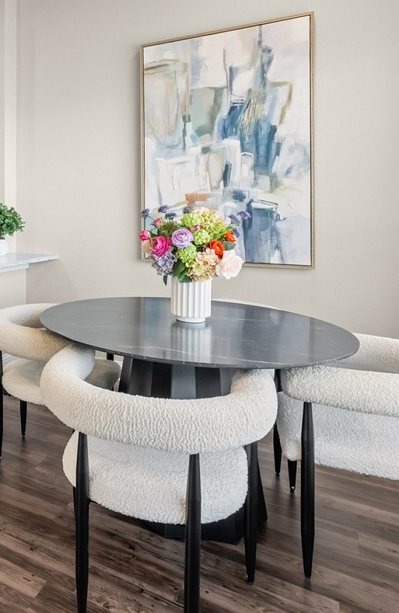 Modern kitchen and dining area with elegant dark cabinetry, stainless steel appliances, and a cozy round table with plush chairs. Bright artwork and vibrant flowers add a touch of sophistication.