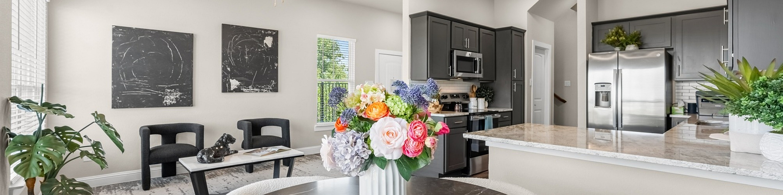 Bright and modern open-concept living area with elegant dining table, vibrant floral centerpiece, sleek kitchen with granite countertops, and contemporary artwork on the walls.