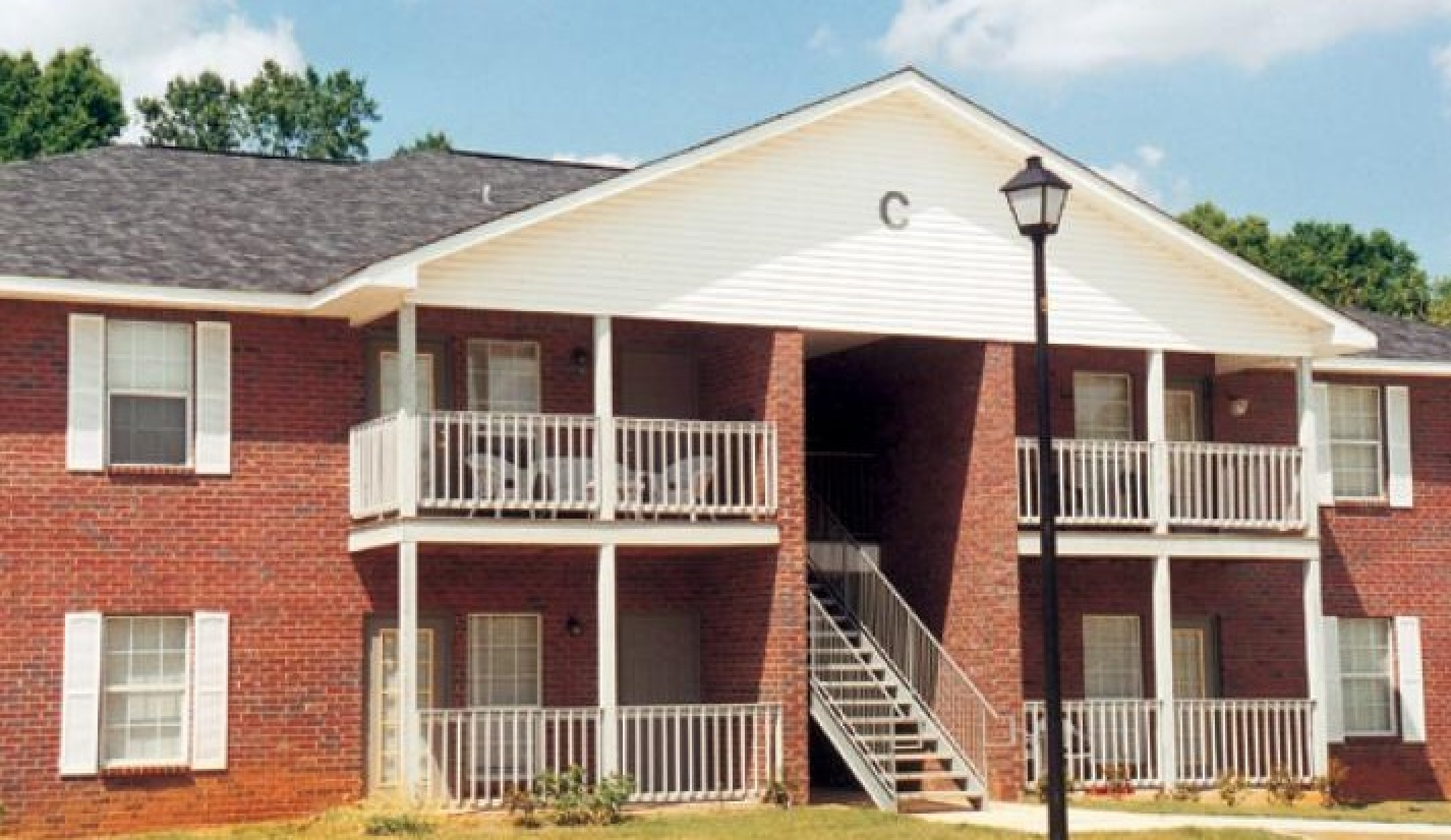 Charming brick apartment building with white trim, featuring covered balconies and nestled beside lush greenery under a vibrant blue sky.