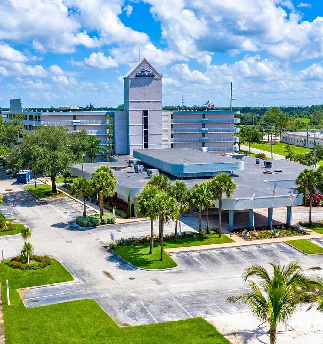 Sunny aerial view of a modern apartment complex with lush landscaping, palm trees, and ample parking, highlighting a vibrant suburban setting with blue skies.