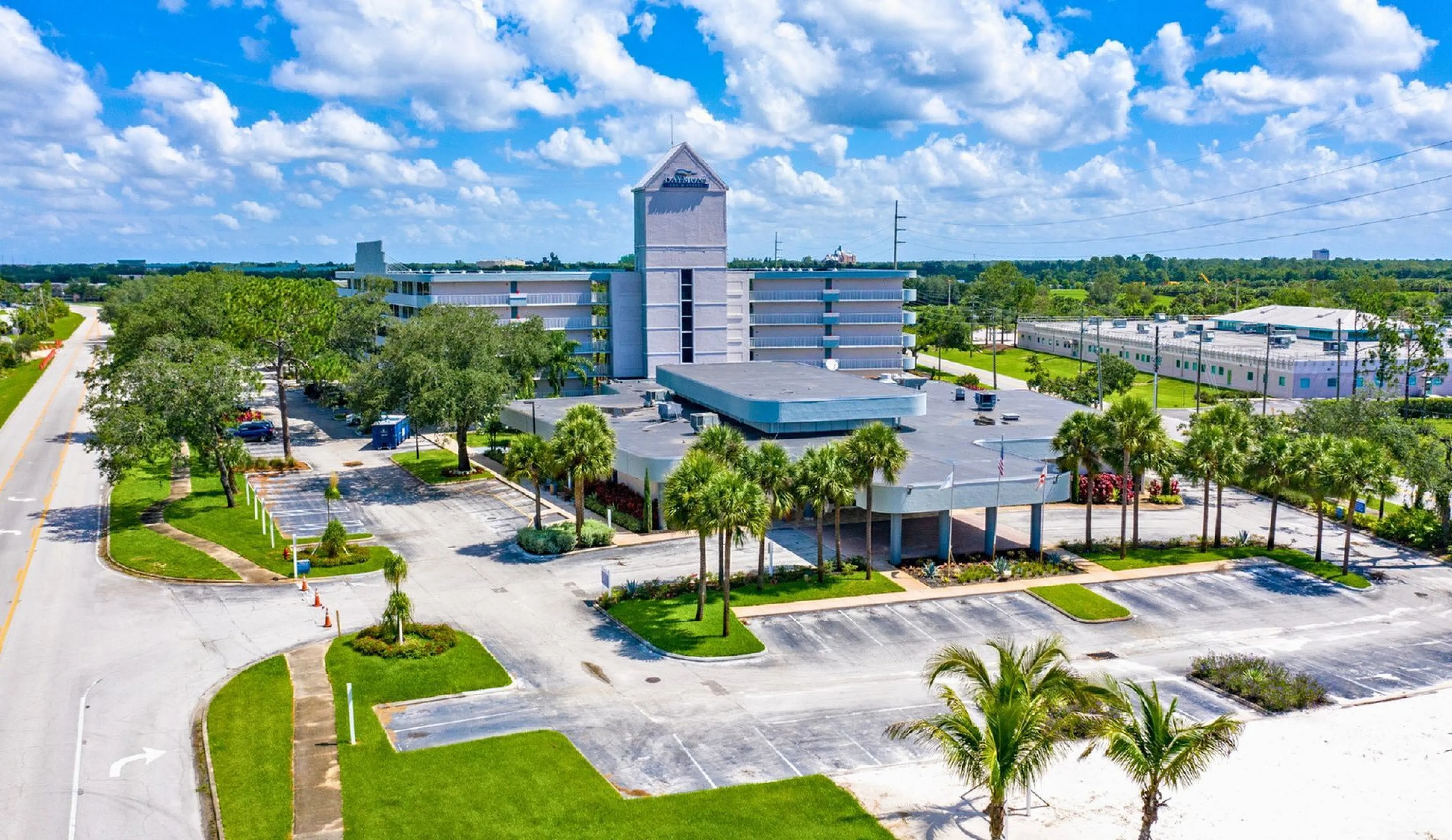 Vibrant aerial view showcasing a modern apartment complex surrounded by lush greenery and clear skies, featuring ample parking and palm tree-lined paths.