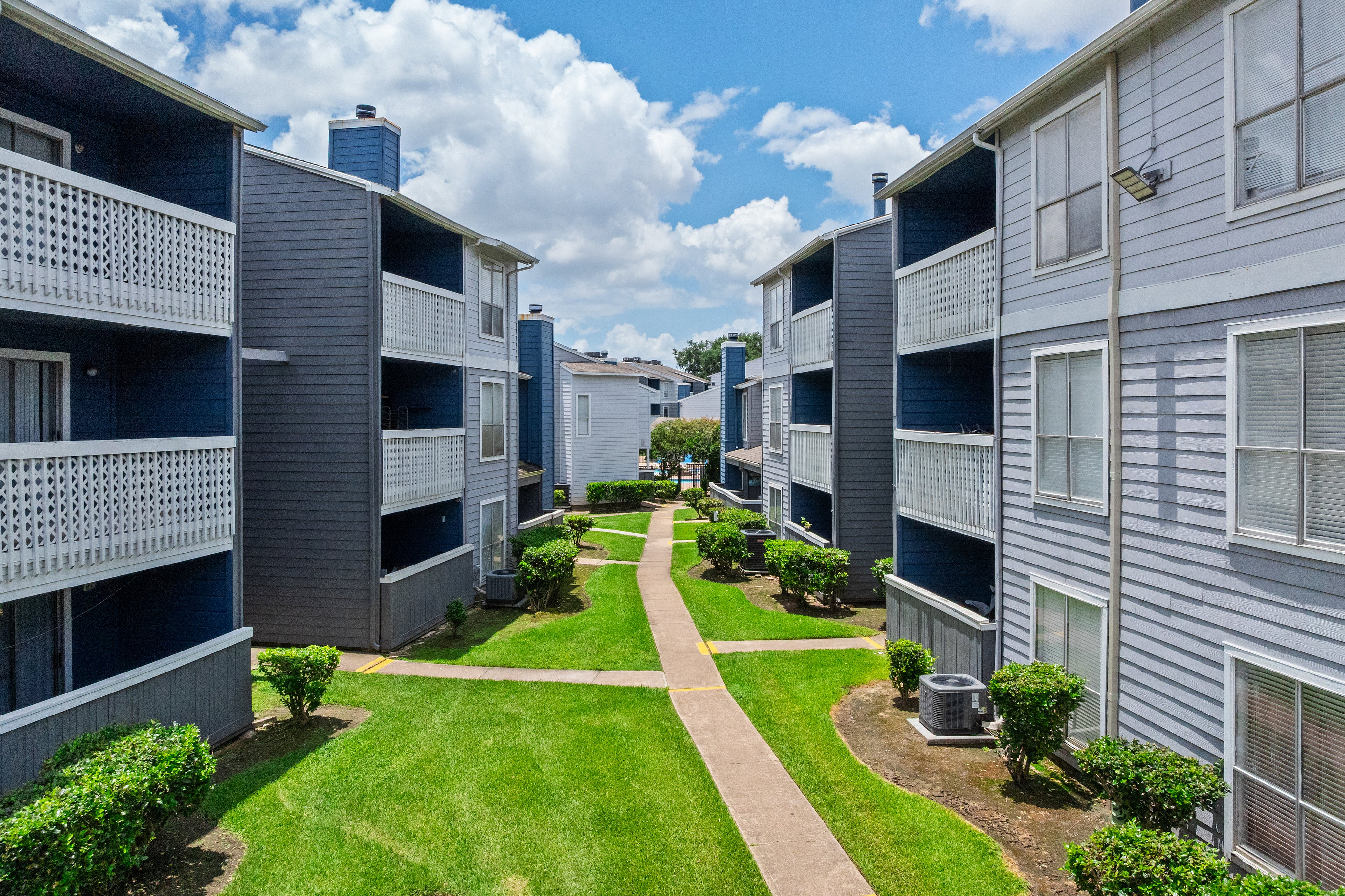 Charming apartment complex with spacious balconies, manicured lawns, and vibrant surroundings under a bright blue sky.
