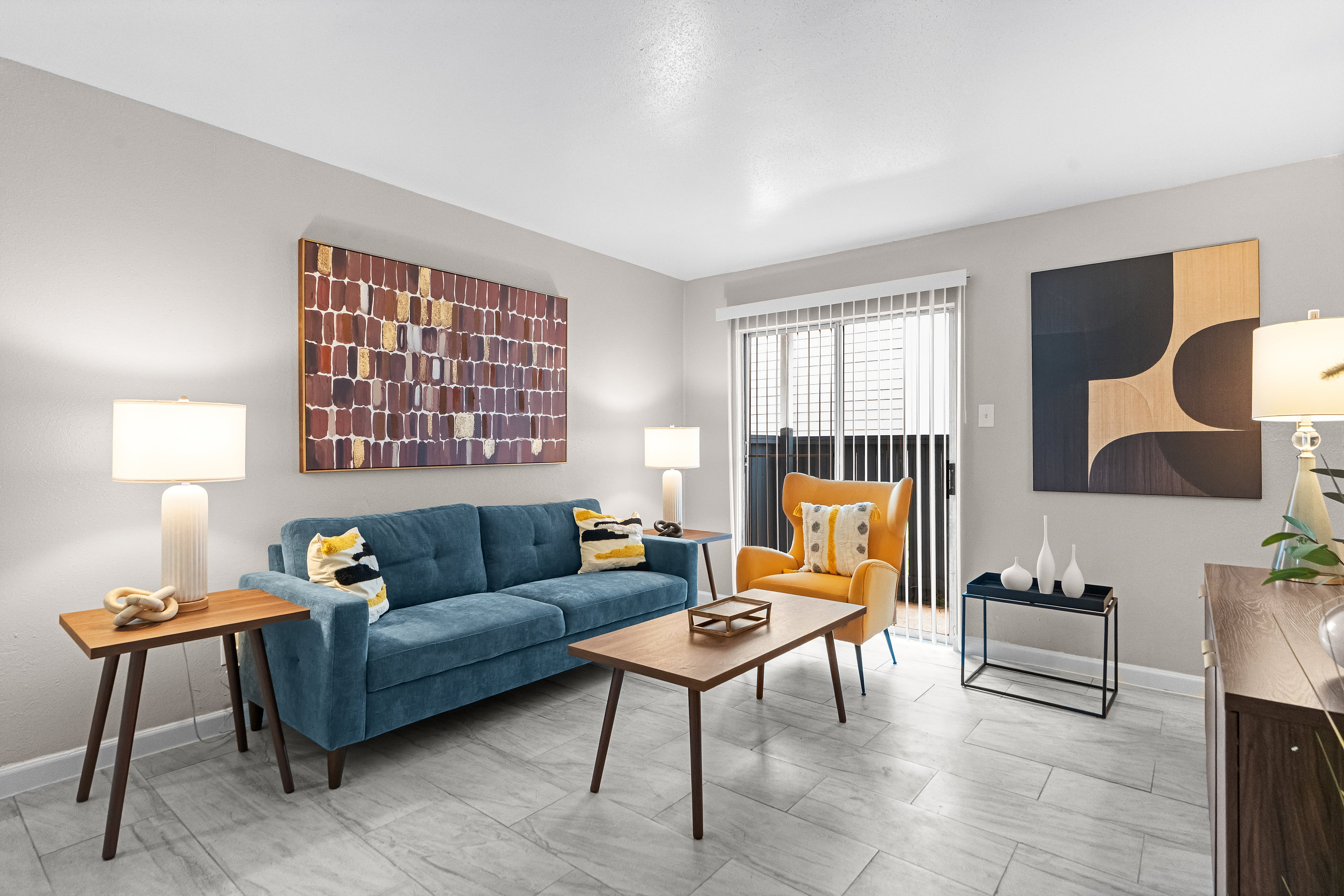Modern living room with a stylish blue sofa, vibrant yellow armchair, and sleek tiled flooring, enhanced by elegant artwork and abundant natural light.