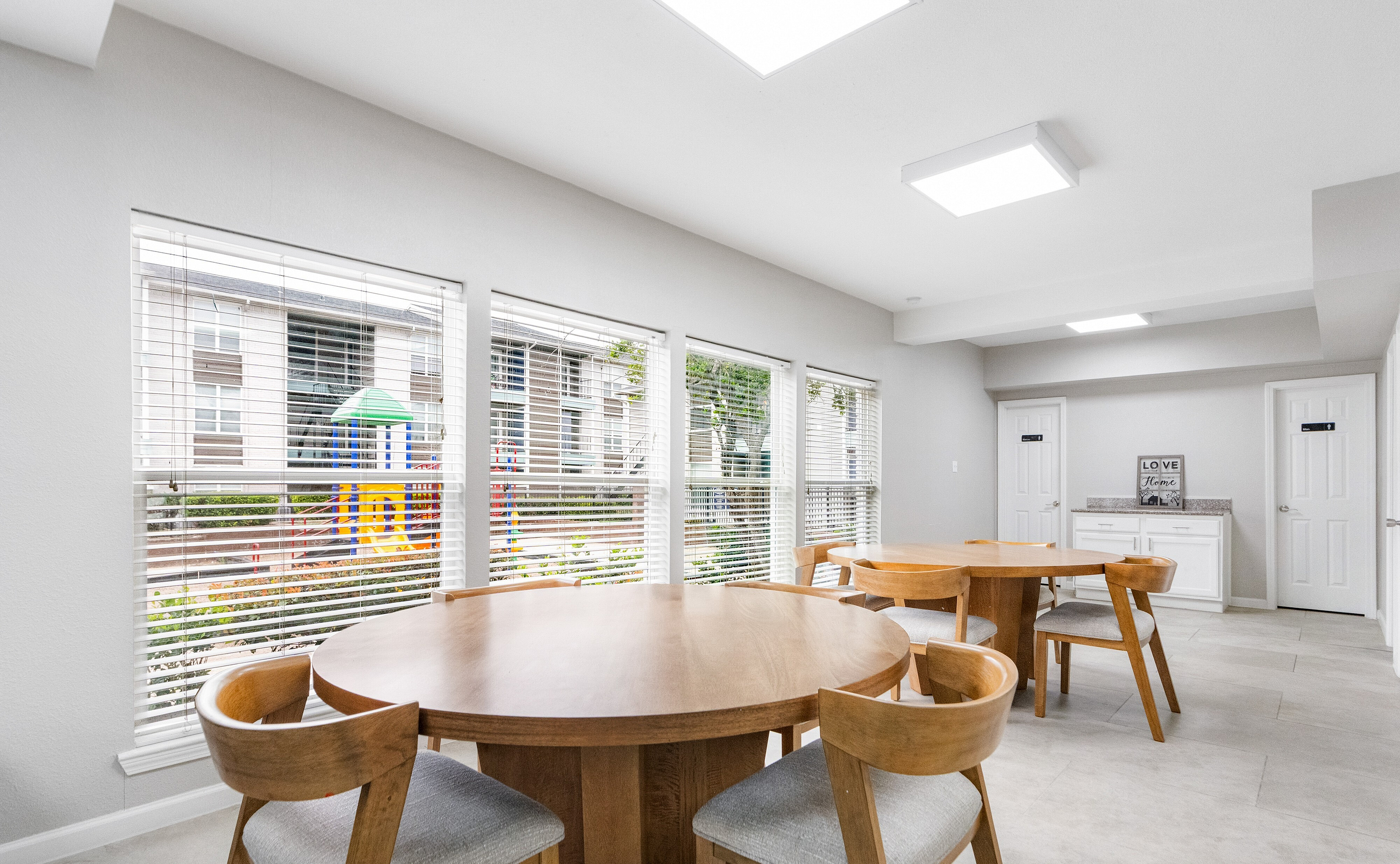 Bright communal dining area featuring elegant wooden tables and chairs with large windows overlooking a playground.