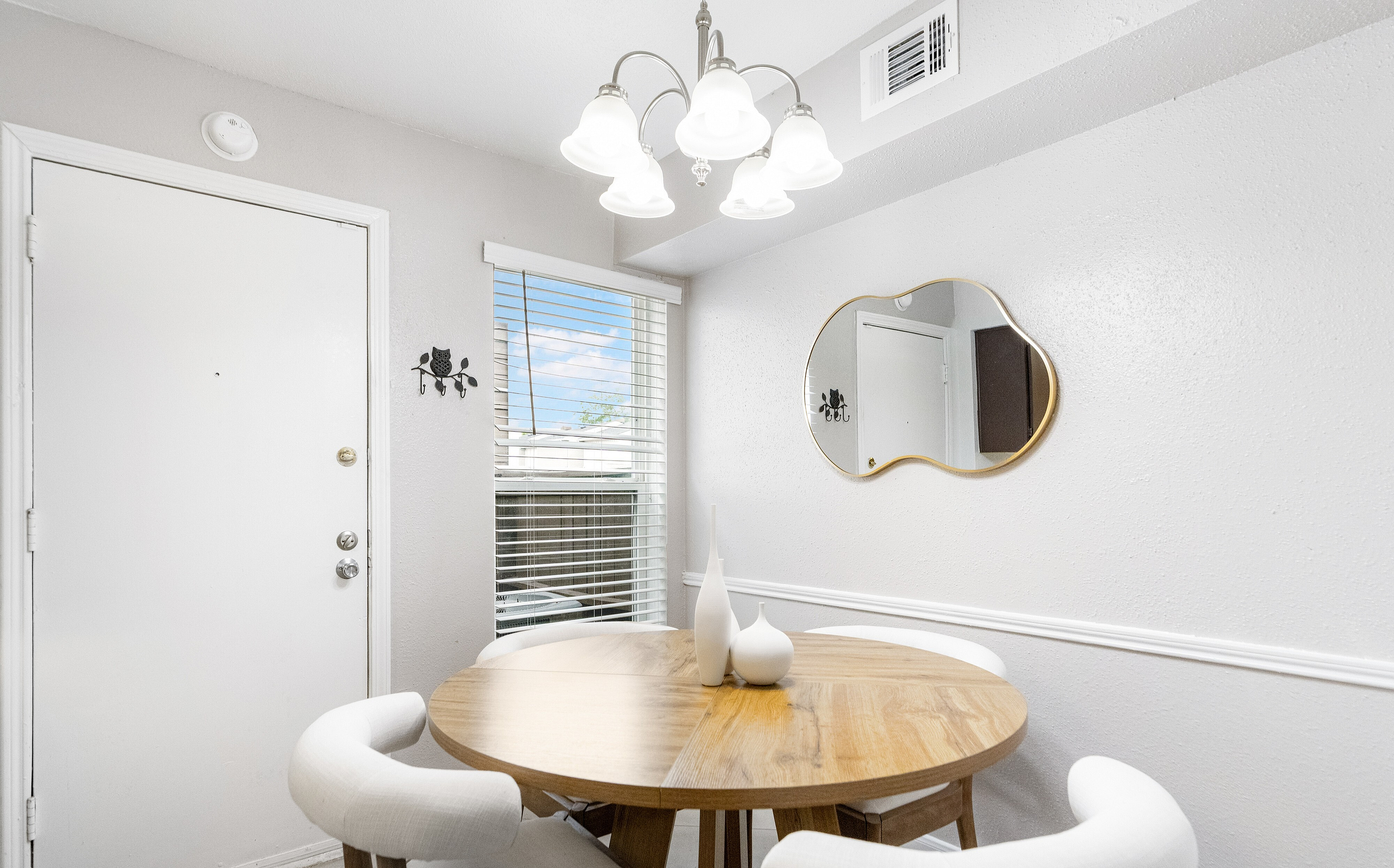 Inviting dining nook with a round wooden table, elegant chandelier, and decorative mirror, bathed in natural light.