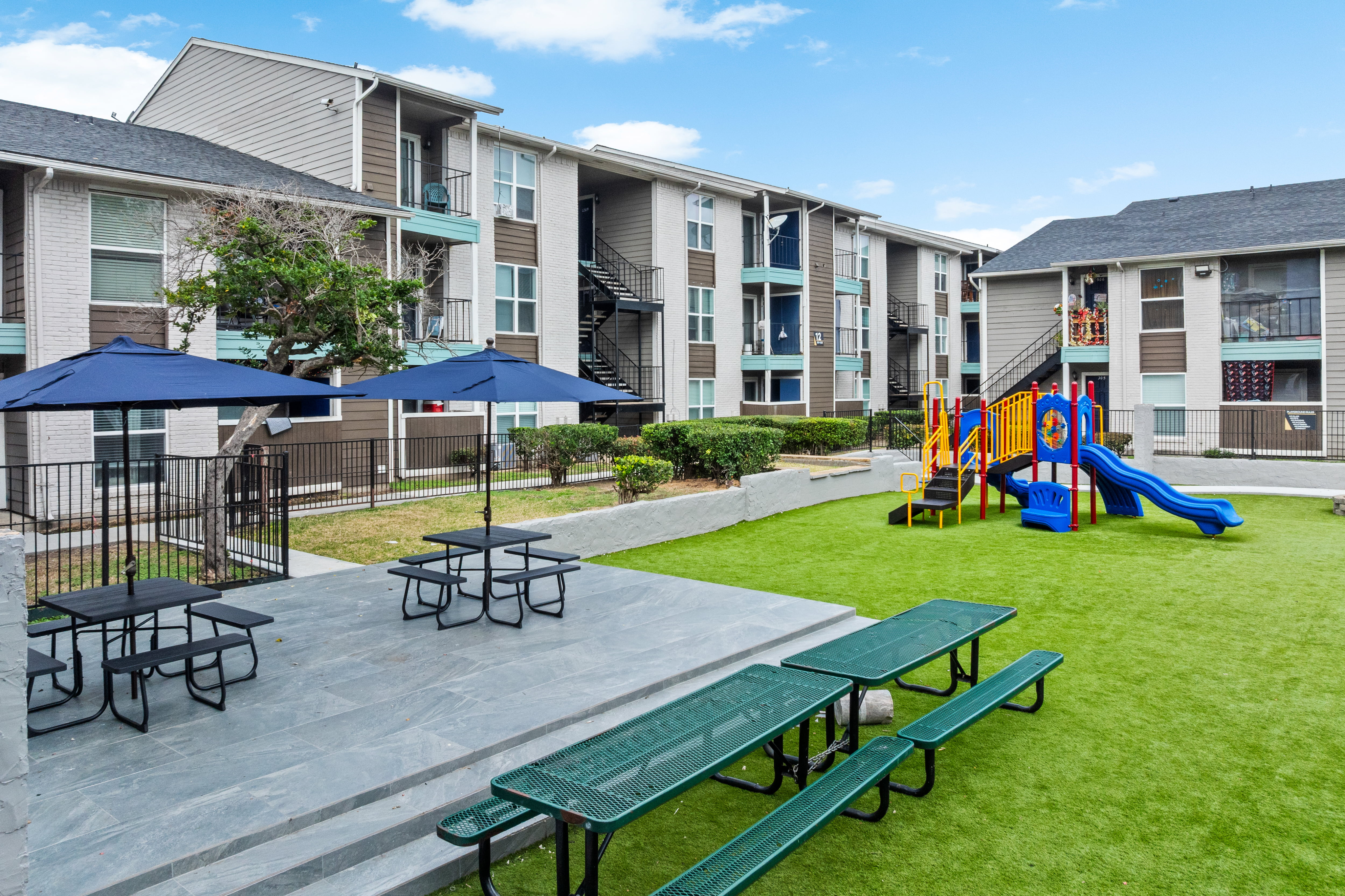 Modern apartment complex featuring communal outdoor space with picnic tables, blue sun umbrellas, and a colorful children's playground set on lush green grass.