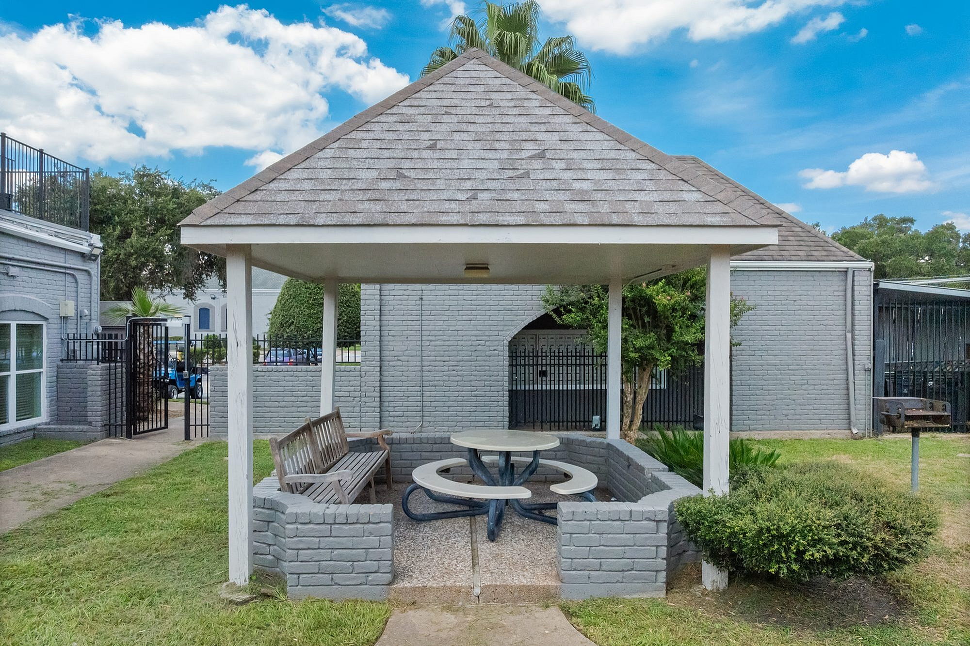 Charming outdoor gazebo with cozy seating and picnic table, surrounded by lush greenery and a bright blue sky.