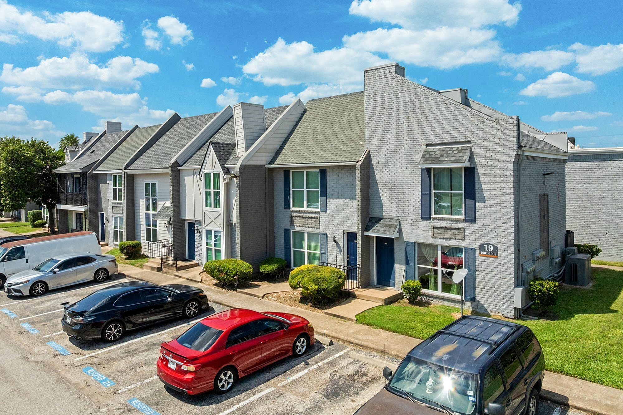 Charming townhouse complex with modern brick facade, lush greenery, and ample parking under a vibrant blue sky.