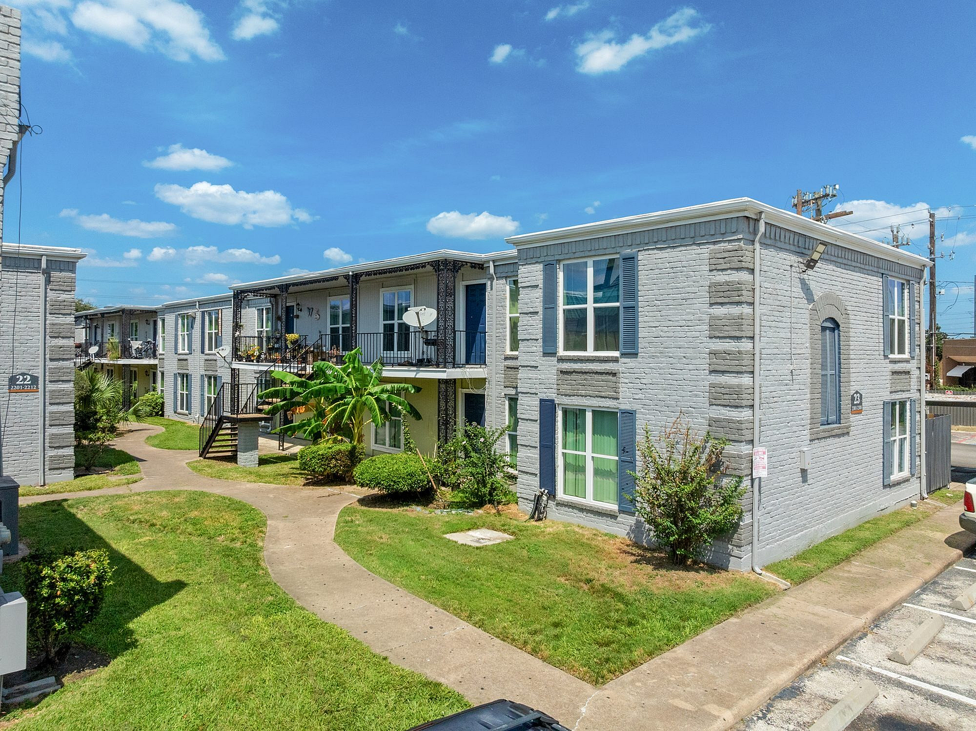Charming apartment complex with gray brick facade, lush greenery, and a clear blue sky, offering a serene and inviting atmosphere.