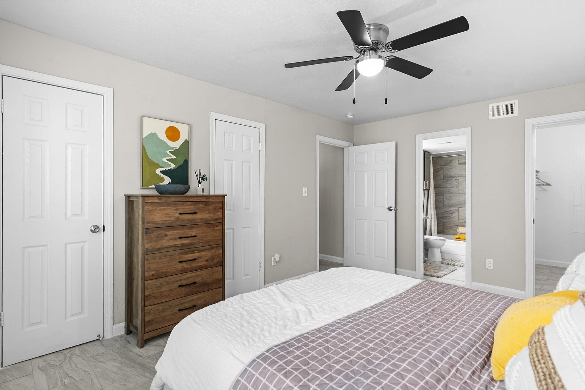 Inviting bedroom with modern ceiling fan, wooden dresser, and tasteful wall art, featuring an ensuite bathroom and ample closet space.