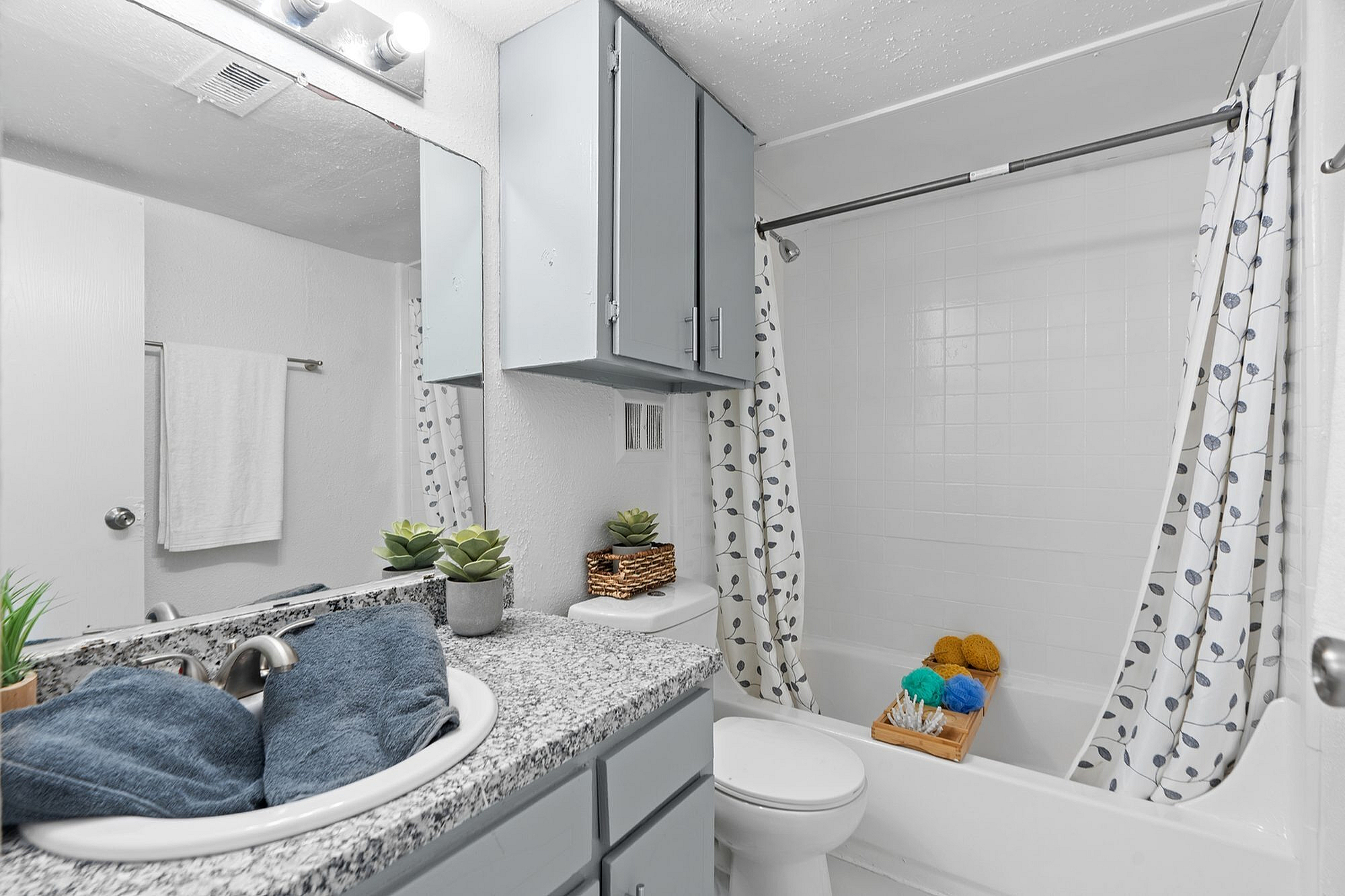 Modern bathroom showcasing elegant granite countertop, sleek fixtures, and minimalist decor. Features a stylish shower with patterned curtain, accentuated by charming plants and neatly arranged essentials. Ideal for relaxation and convenience.