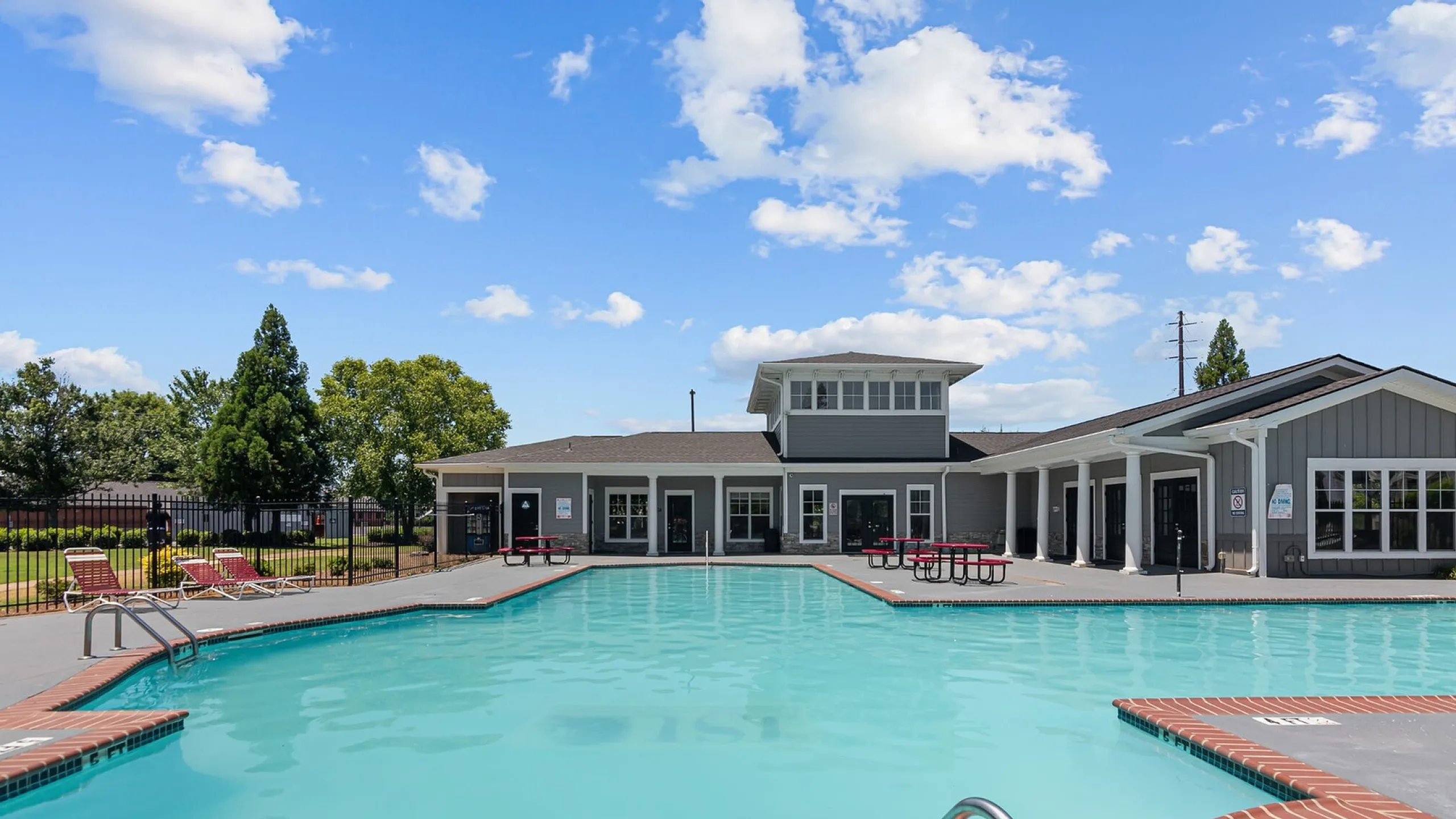 Resort-style swimming pool with modern clubhouse, surrounded by lush greenery and sunny skies, featuring comfortable loungers and picnic tables.