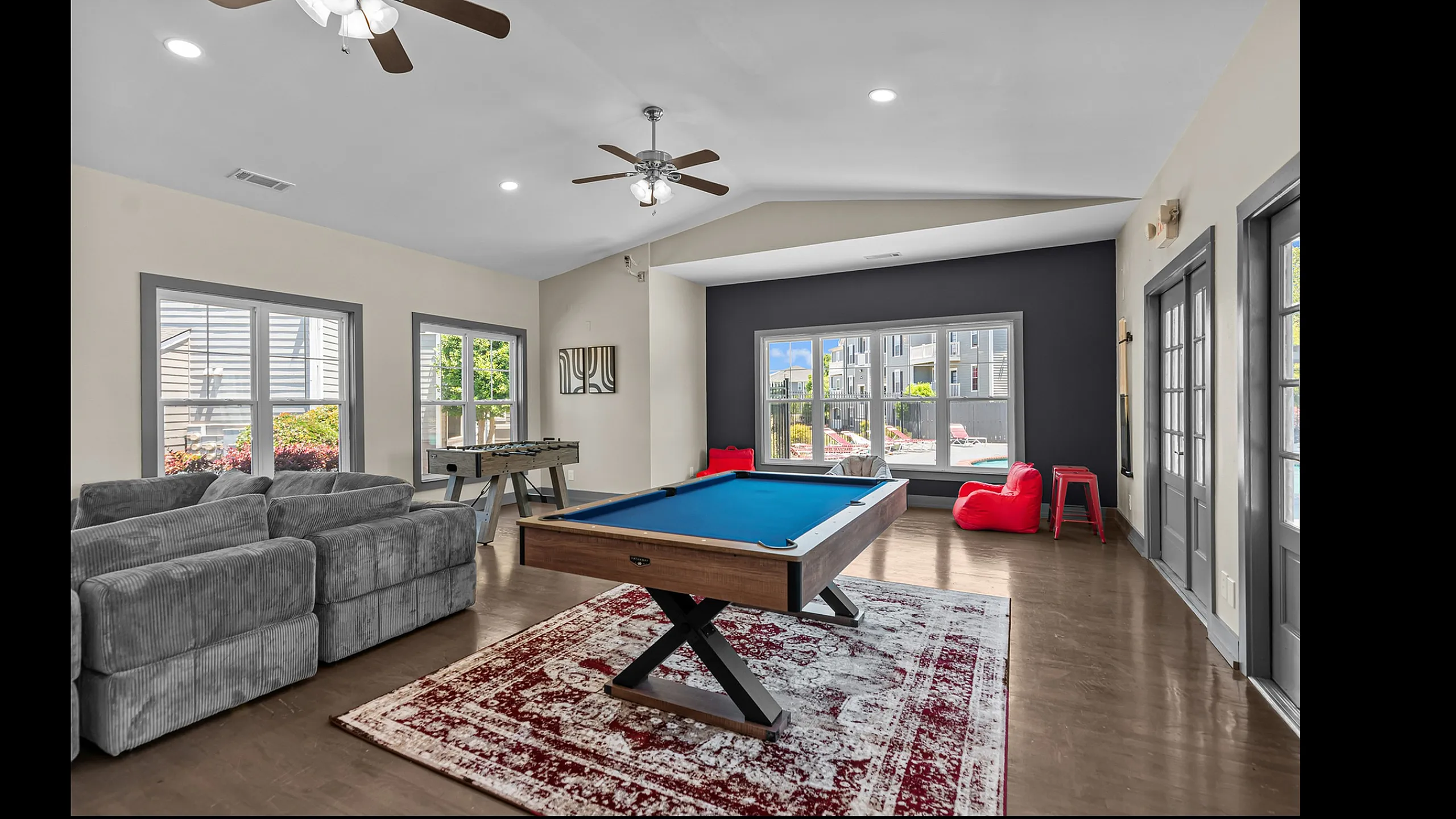 Chic game room featuring a stylish pool table, cozy grey sofas, and a vibrant area rug. Bright windows provide a stunning view of the outdoor pool area.