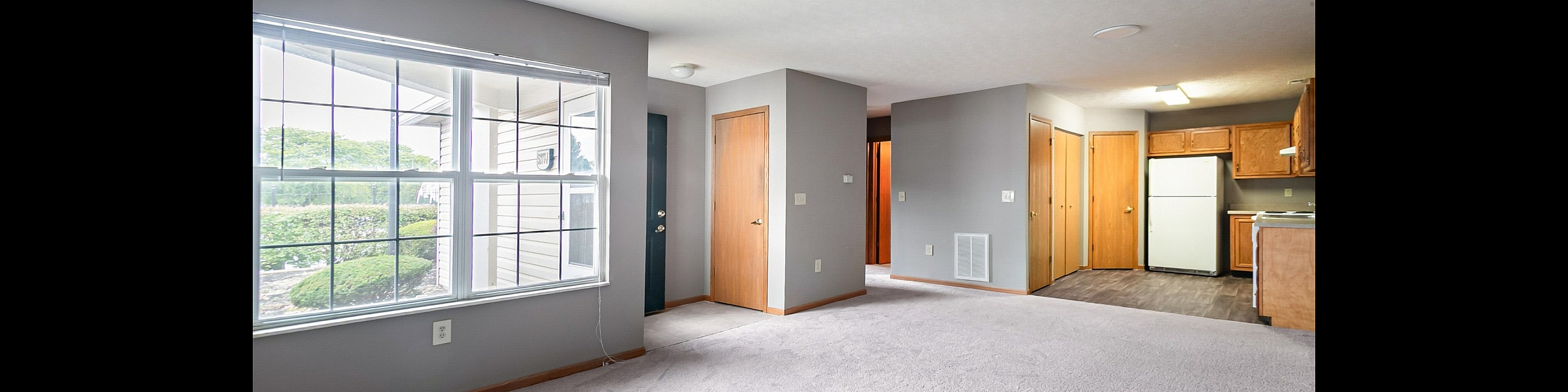 Inviting apartment interior with large windows allowing natural light, neutral carpeted flooring, and a cozy kitchen area.