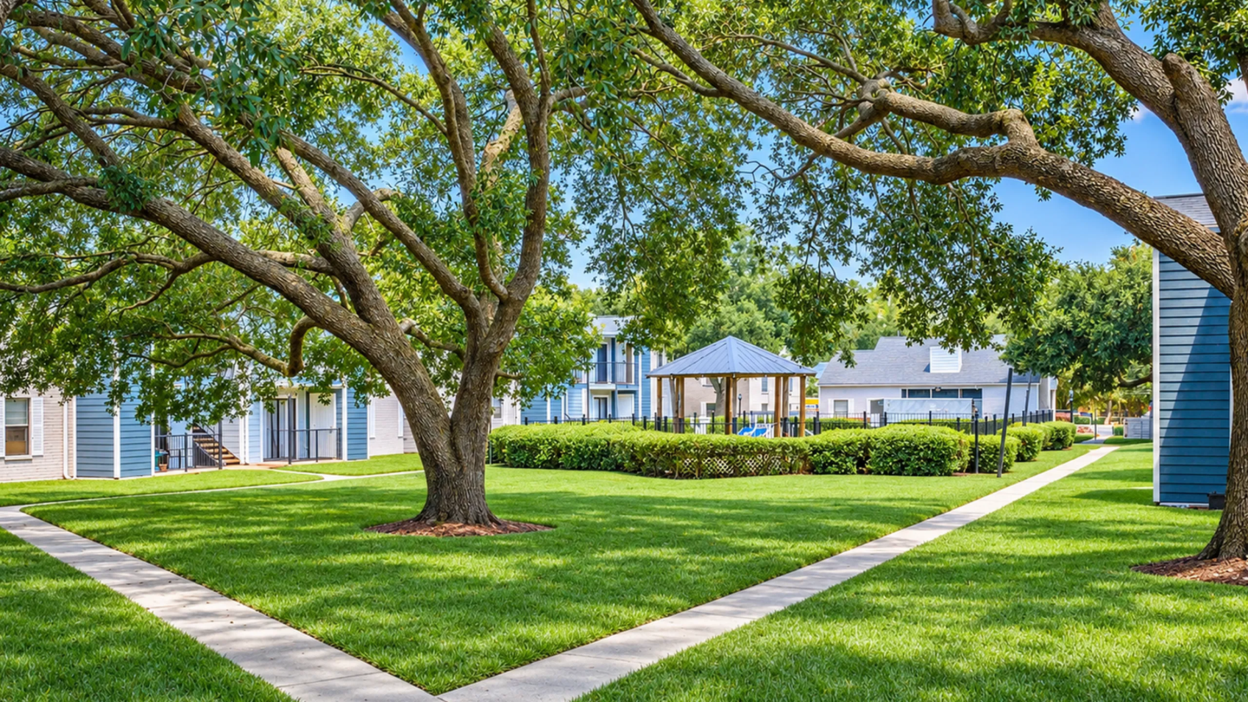 Serene apartment courtyard with lush greenery, shaded by mature trees, and a charming gazebo offering a peaceful retreat.