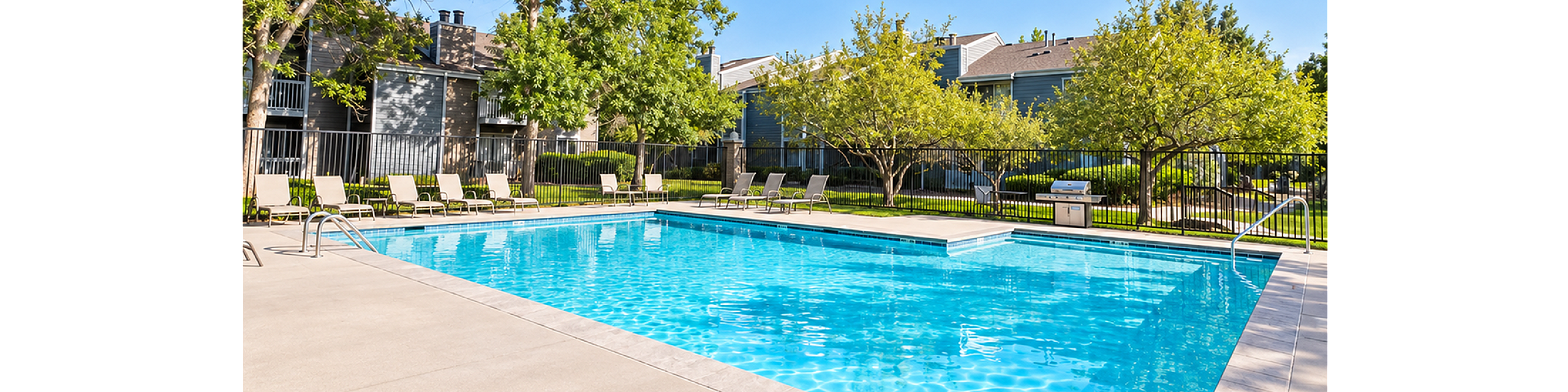 Inviting outdoor pool area with sun loungers, surrounded by lush greenery and modern apartment buildings under a clear blue sky.