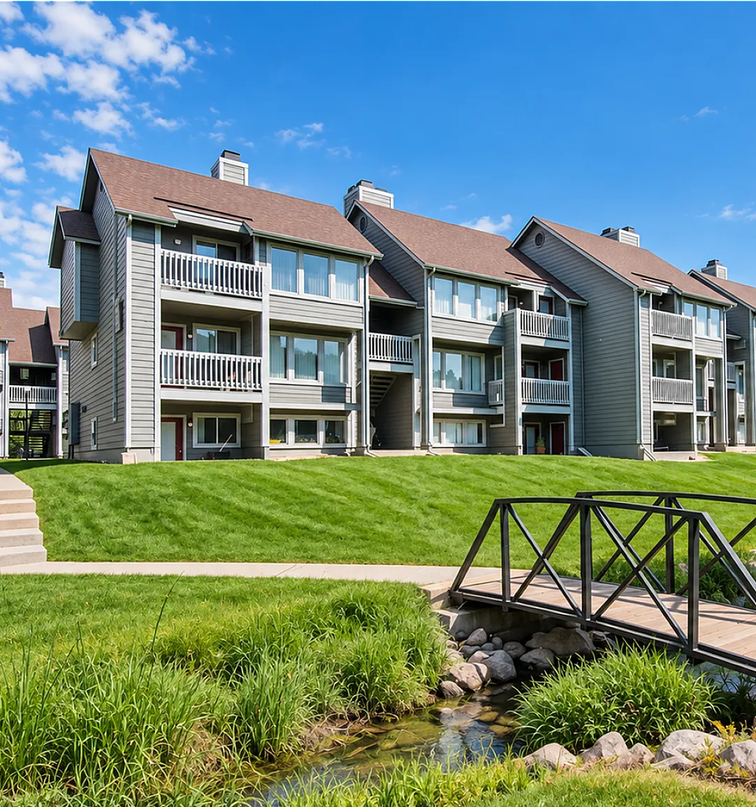 Charming apartment complex with modern architecture, lush green landscaping, and a scenic walking bridge under a vibrant blue sky.