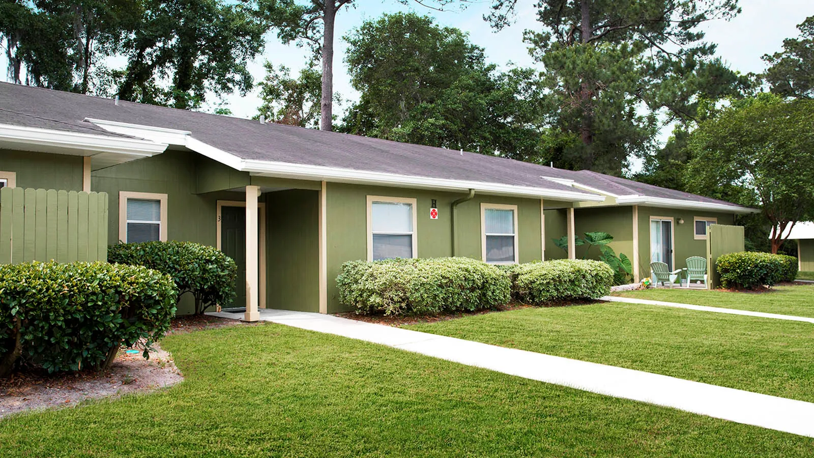 Apartment building painted light green with green grass, lush landscaping and trees in the background.