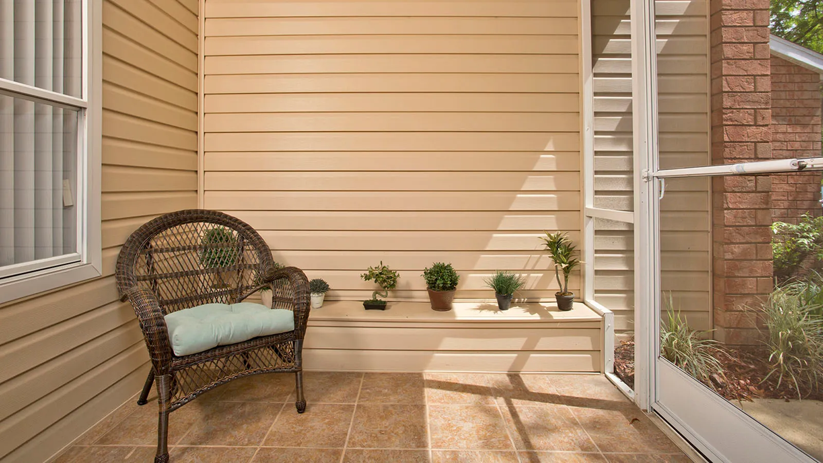 Screened patio with tile floor and wicker chair.  Small plants sitting on top of a built in bench along the wall.