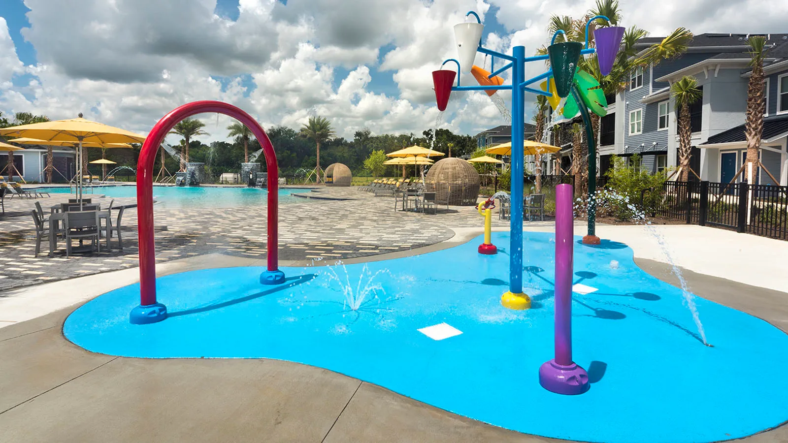 Children's splash pad with various shapes and fountains.  Community pool, beach umbrellas, buildings and trees in background.
