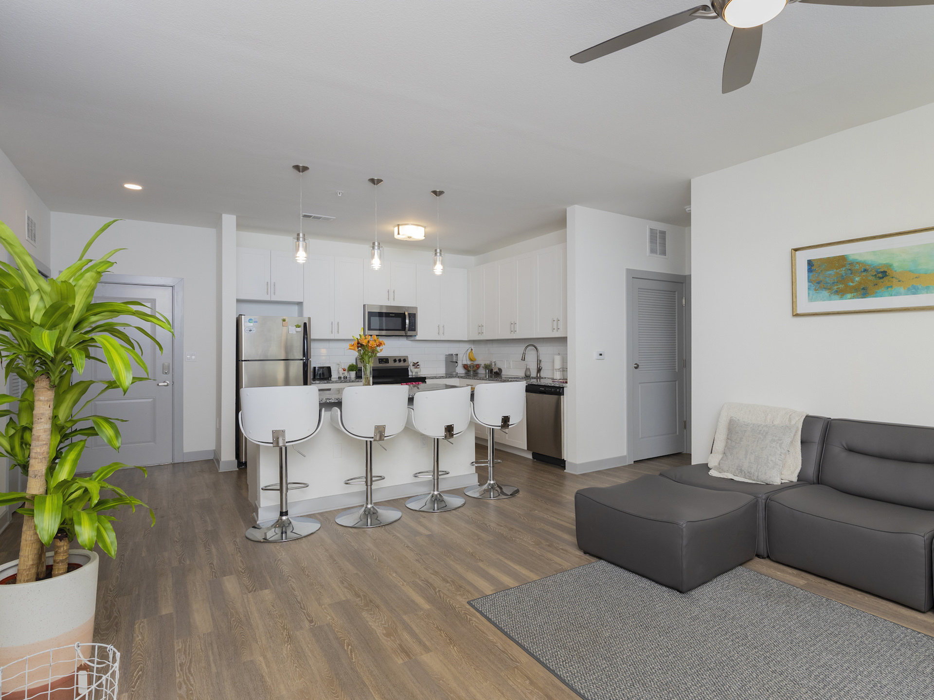 Living room with hardwood floor, area rug, couch, kitchen with island, stainless steel appliances, and white cabinets.