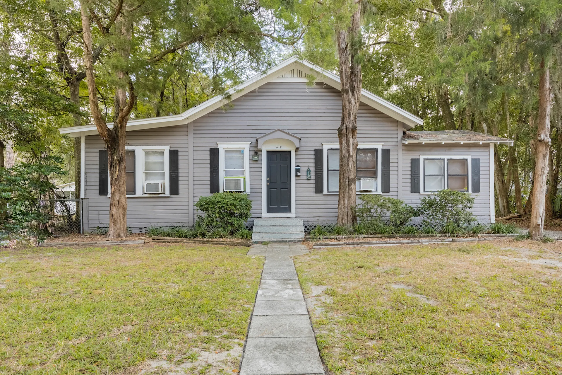 Exterior of the 1 story Willow House. There is a sidewalk leading up to the front steps, and the house has grey sidings, blue door, and green space out front.