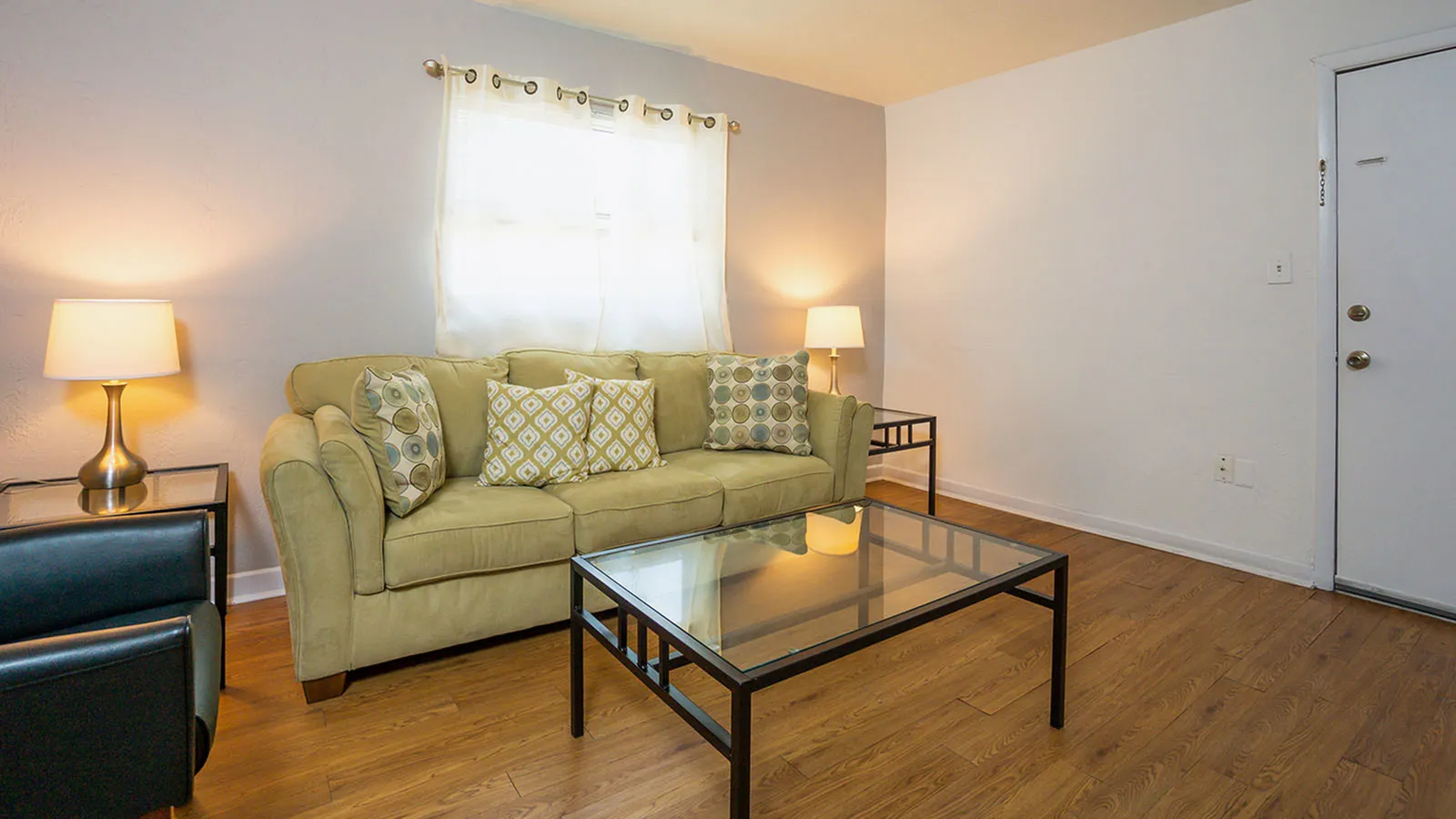 Living area with wood floor, light green fabric covered couch, glass coffee and end tables with lamps.