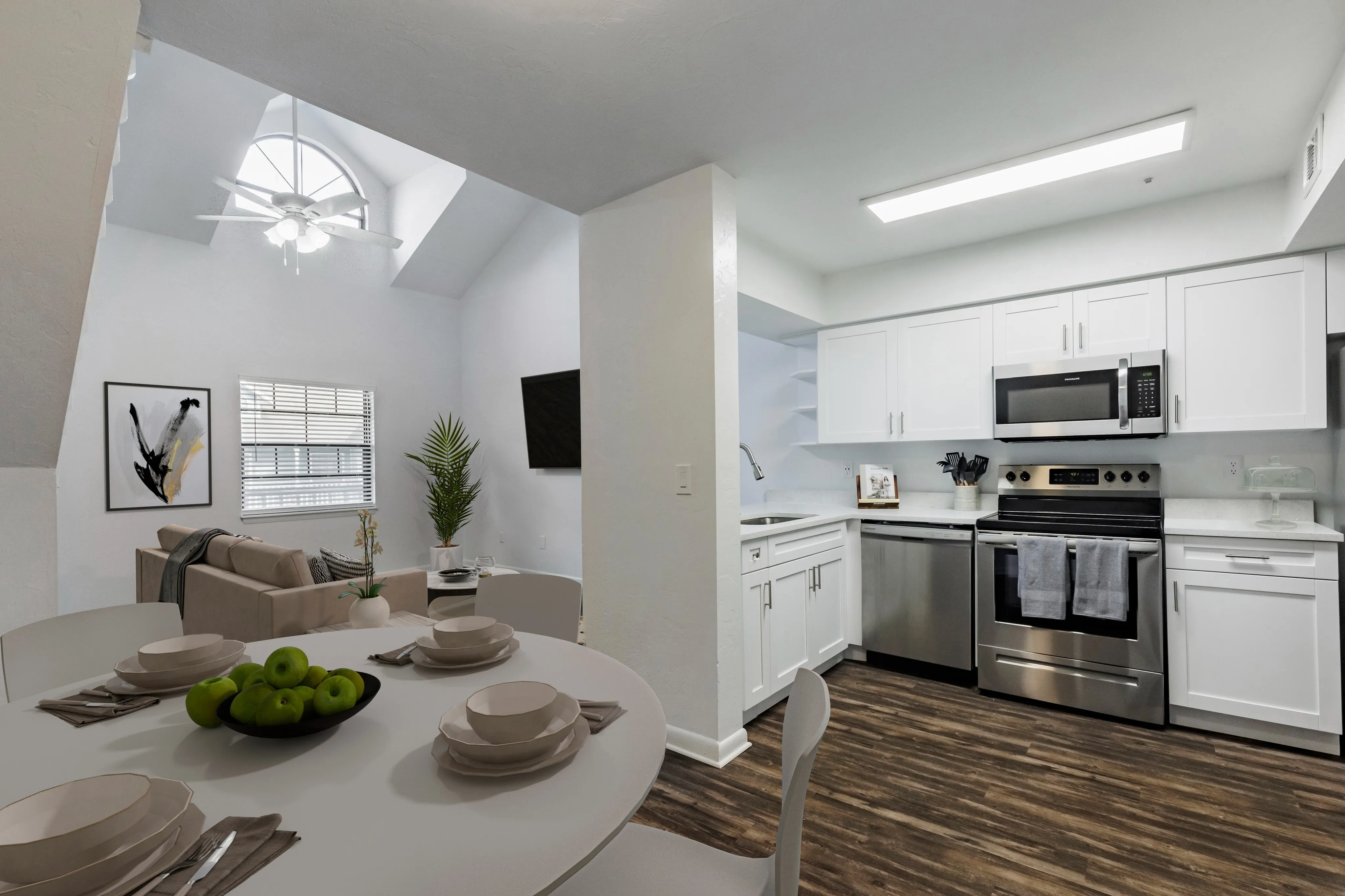 Kitchen with wood floors, premium appliances, dining table in foreground. Background is living room with vaulted ceiling, couch and TV