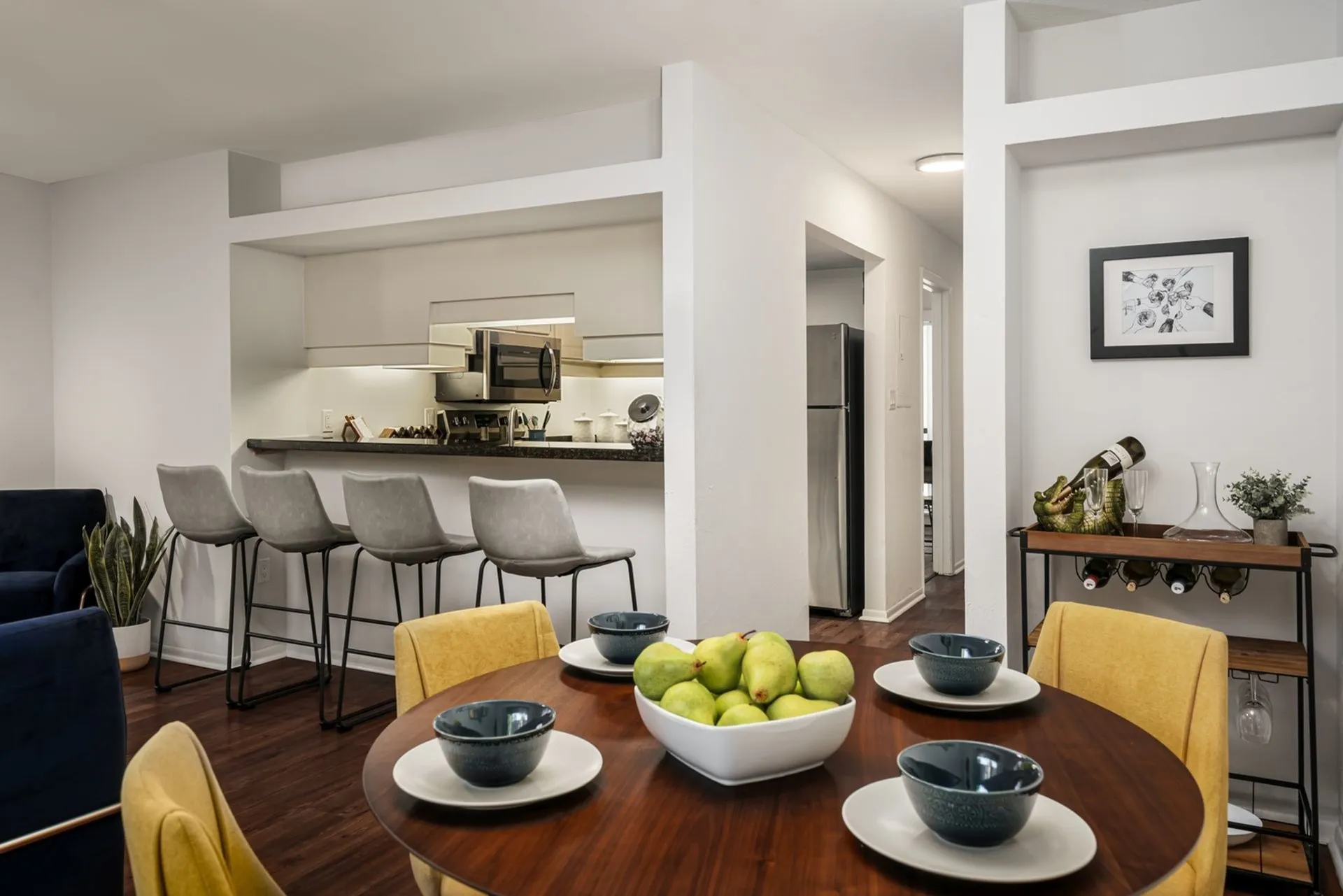 Dining area with wooden table and yellow chairs. Kitchen and breakfast bar visible in the background.