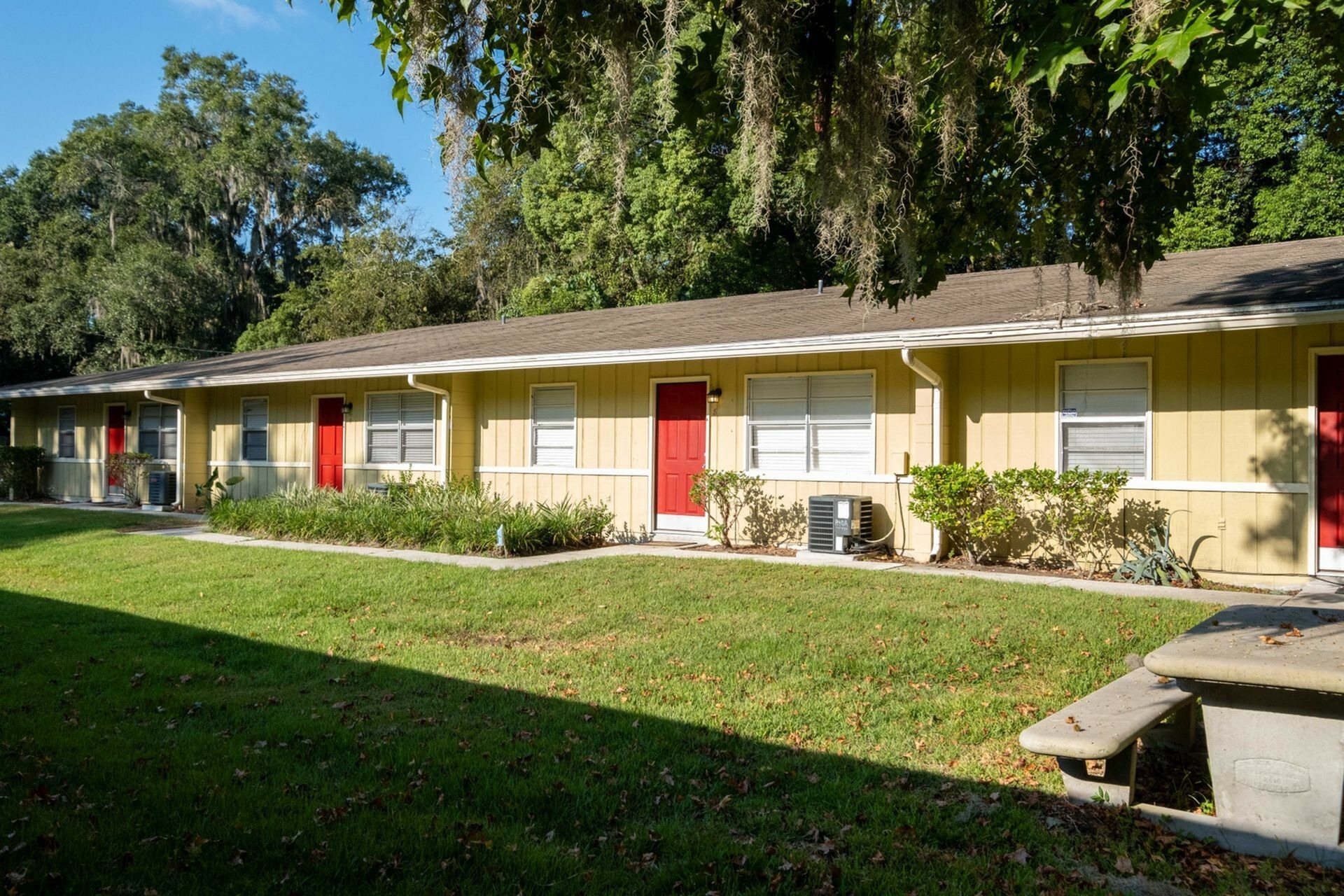 Facade of one level apartment building showing several front doors behind lush green grass and landscaping.
