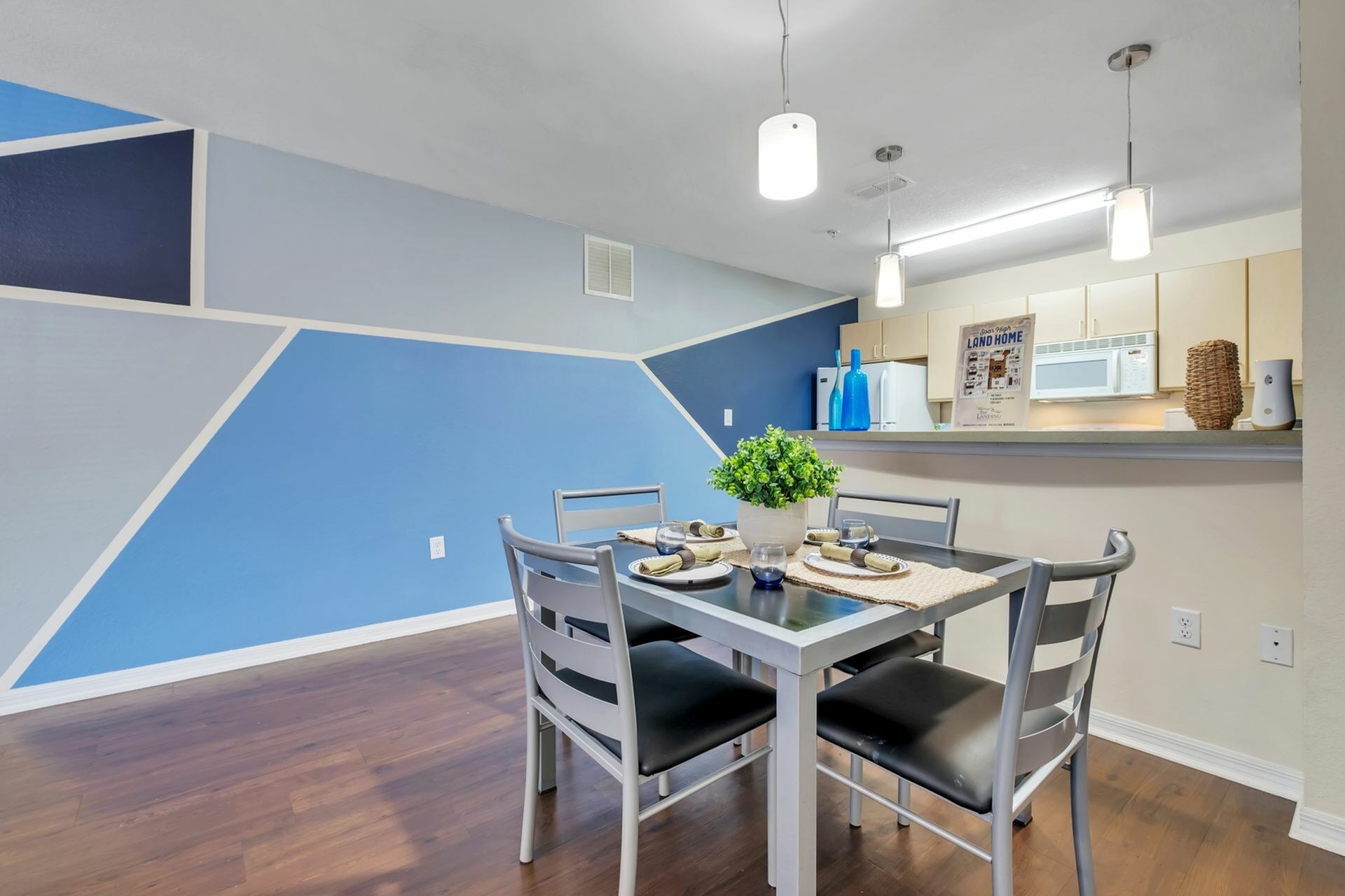 Wide shot of the dining area with a square table and chairs, and living area in the background with a couch. There are hardwood floors and white and blue walls.