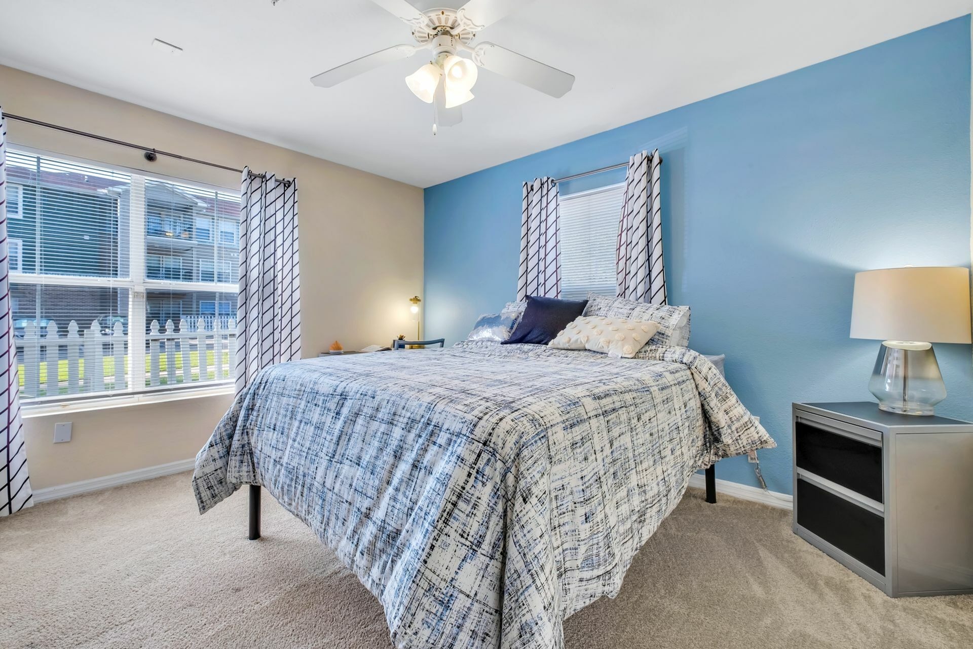 Carpeted bedroom with blue accent wall, desk, bed, night stand and a large window to the left.