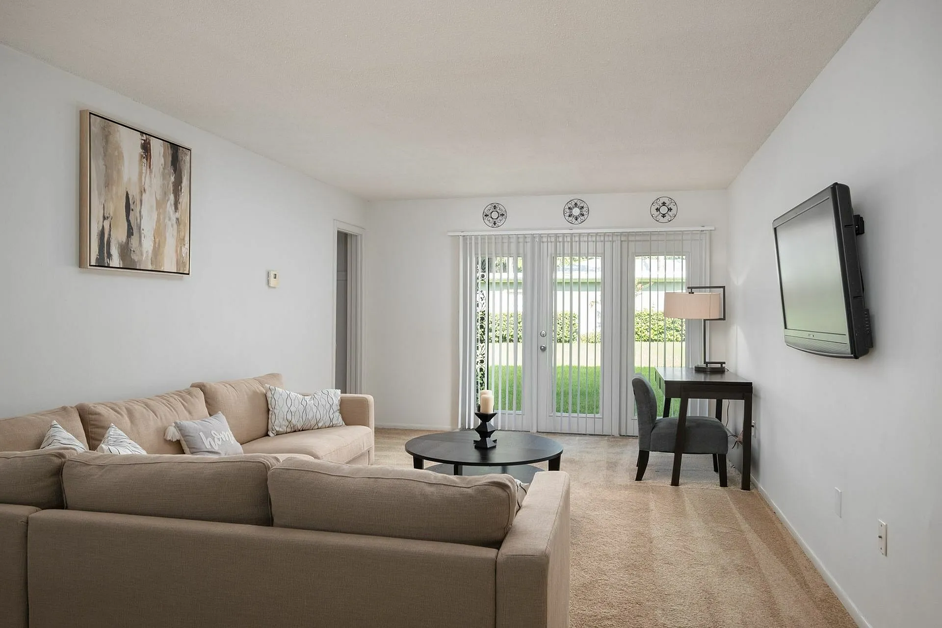 Bright living room with beige sofa, wall-mounted TV, and patio doors leading to garden.