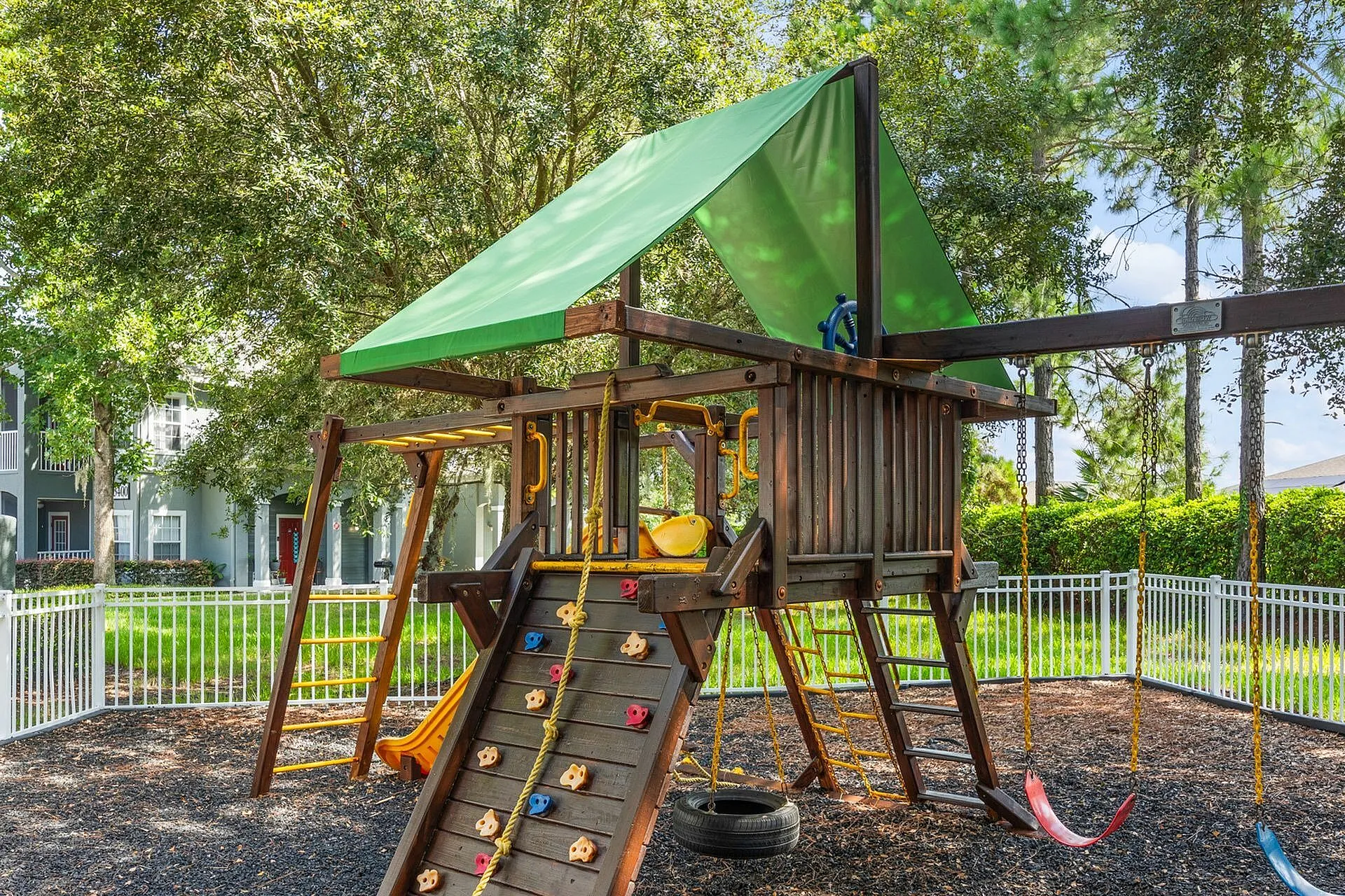 Charming outdoor playground with climbing wall, swings, slide, and wooden playhouse surrounded by lush greenery.