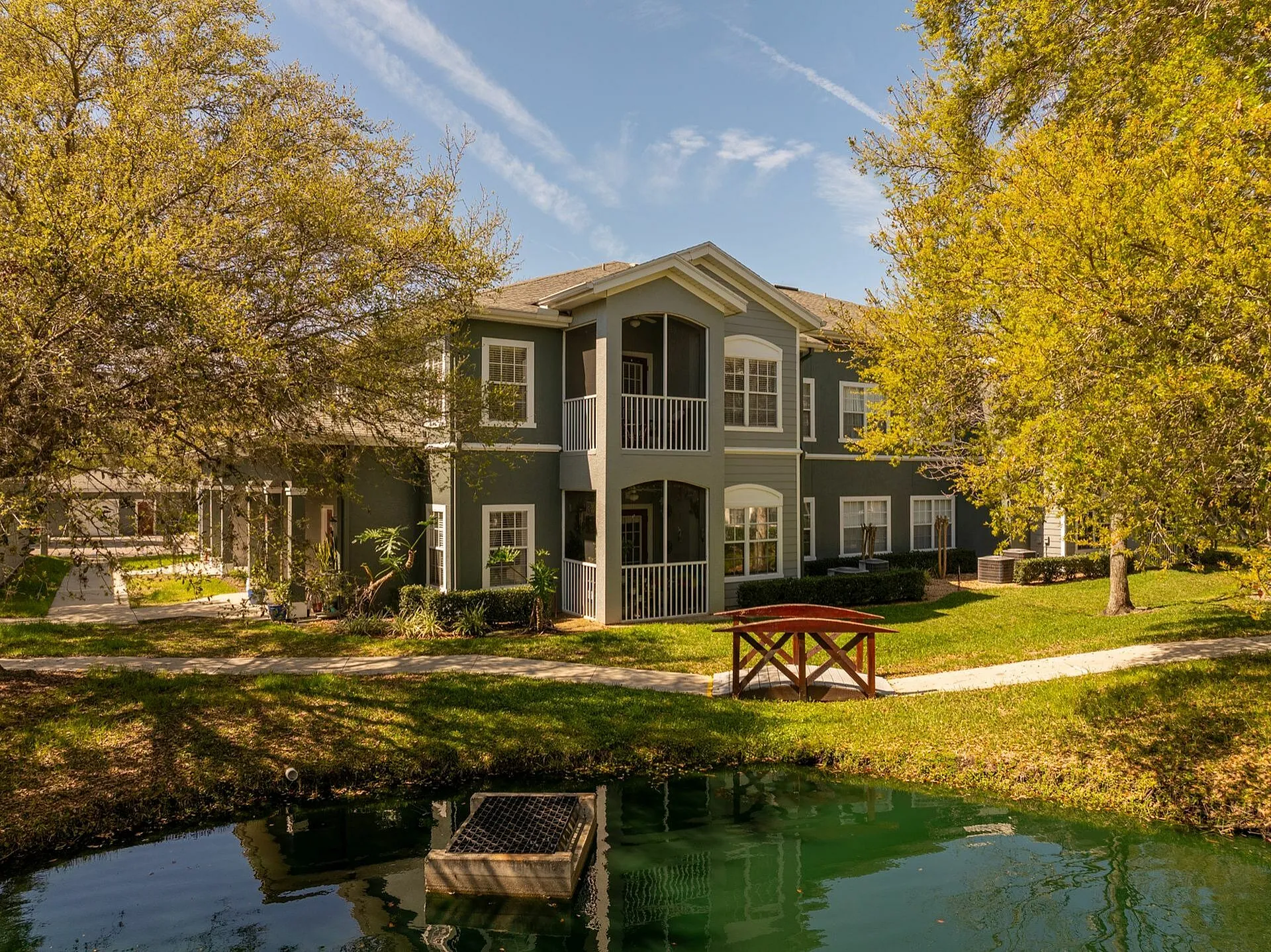 Charming apartment building surrounded by lush greenery and serene pond, featuring a quaint wooden bridge and inviting balconies.