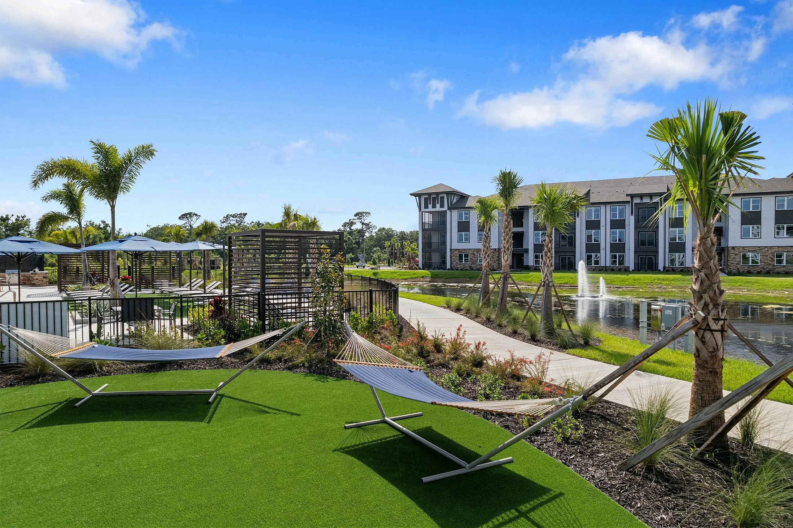 Vibrant apartment complex featuring palm-lined pathways, relaxing hammocks, and peaceful water feature under a bright blue sky.