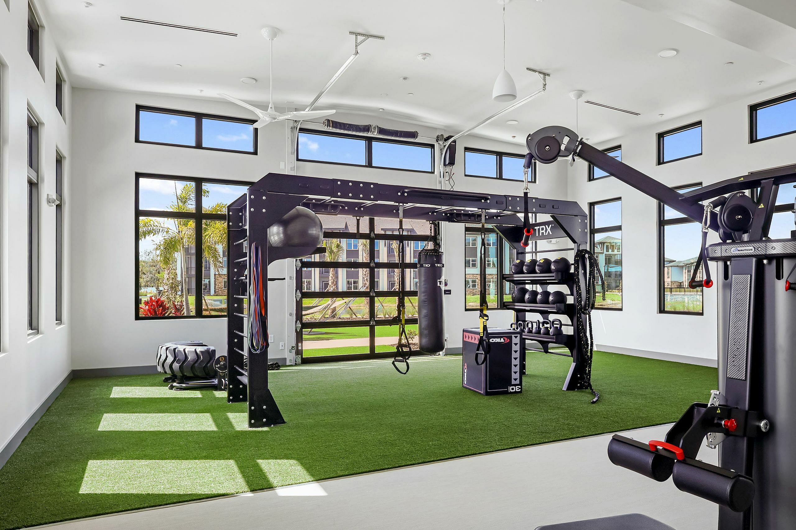 Bright and modern fitness room with state-of-the-art equipment, featuring TRX system, kettlebells, and vibrant greenery outside large windows.