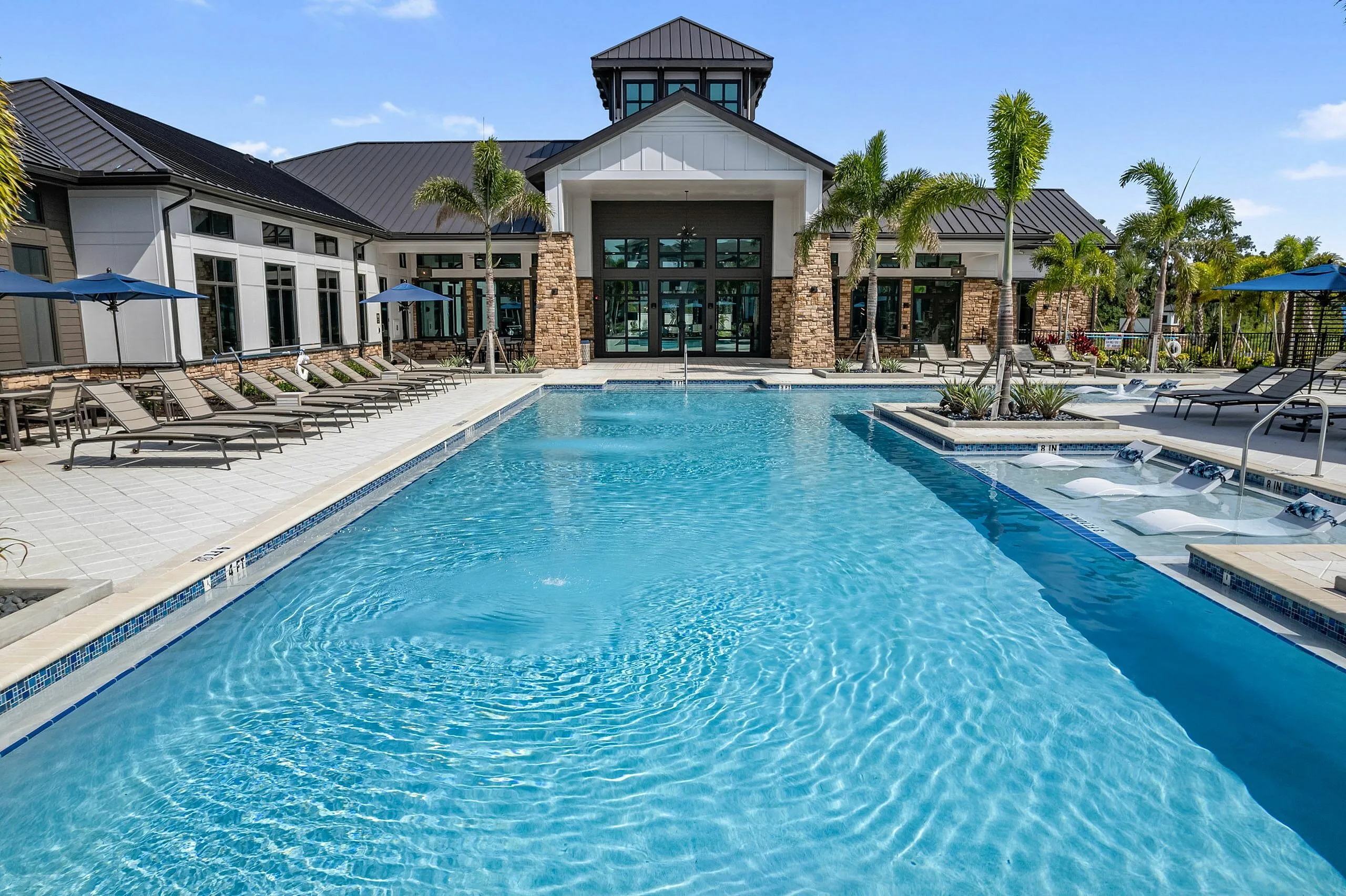 Luxurious poolside oasis with comfortable loungers and modern clubhouse surrounded by swaying palms under a clear blue sky.