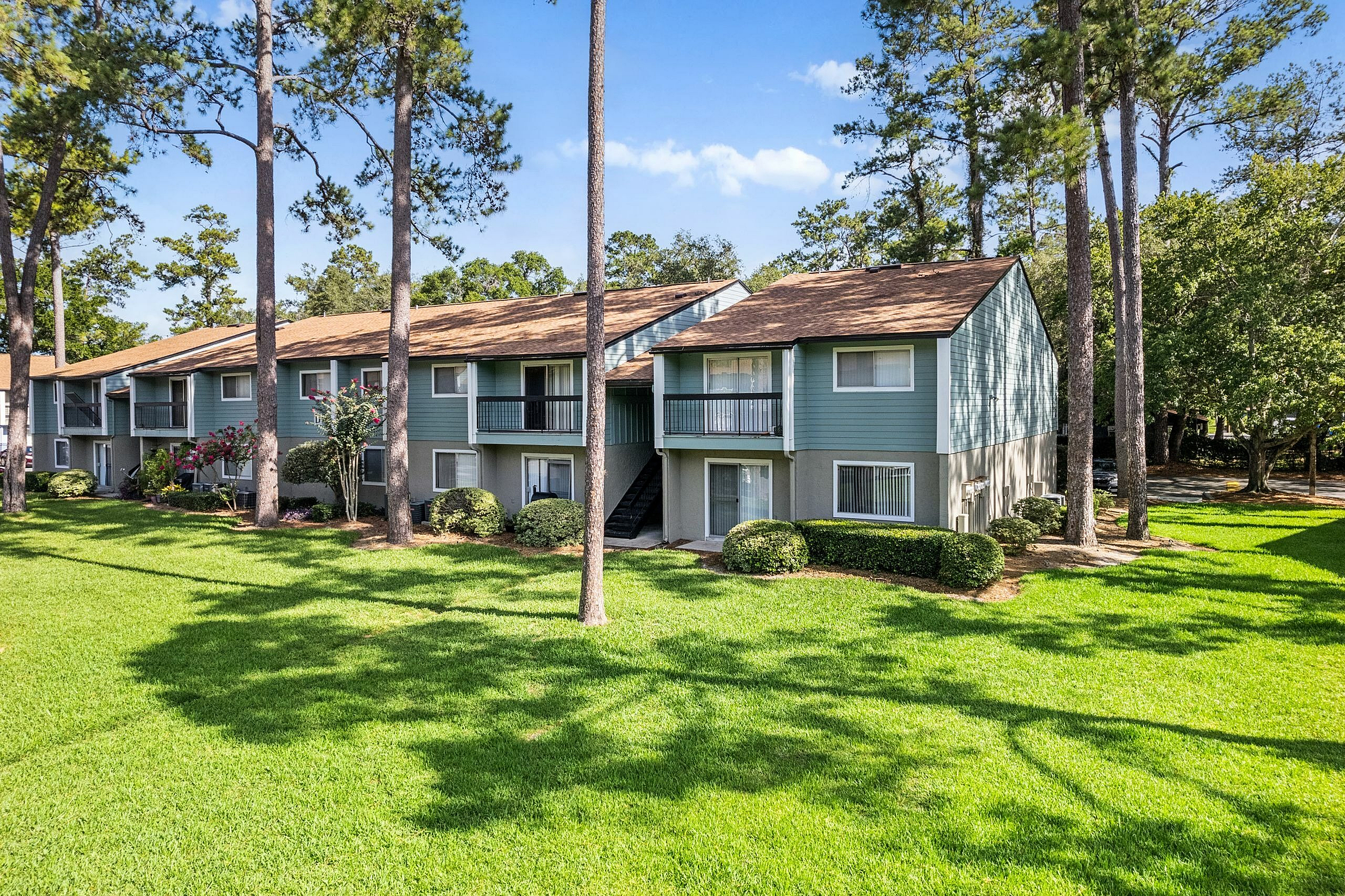 Charming two-story apartment building with a beautifully landscaped lawn, surrounded by tall pine trees and lush greenery, under a bright blue sky.