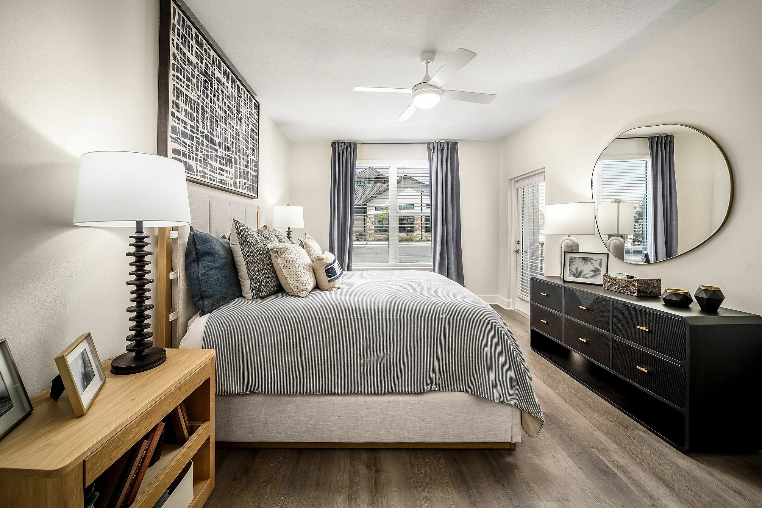 Elegant bedroom featuring a cozy bed with stylish pillows, large window with gray curtains, modern ceiling fan, and sleek black dresser with round mirror.