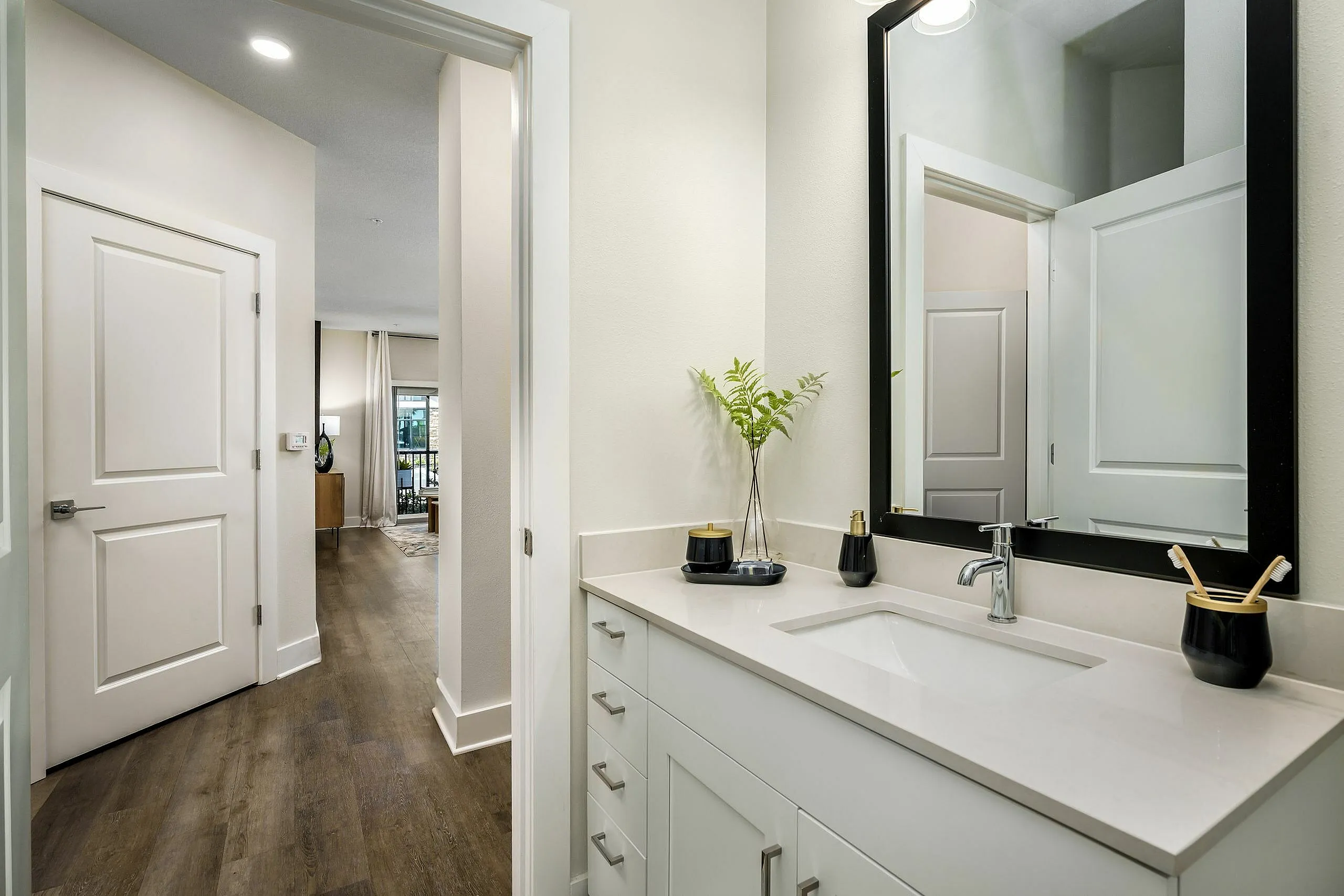 Bright and modern bathroom with elegant vanity, framed mirror, and sleek wood flooring leading to a cozy living space.