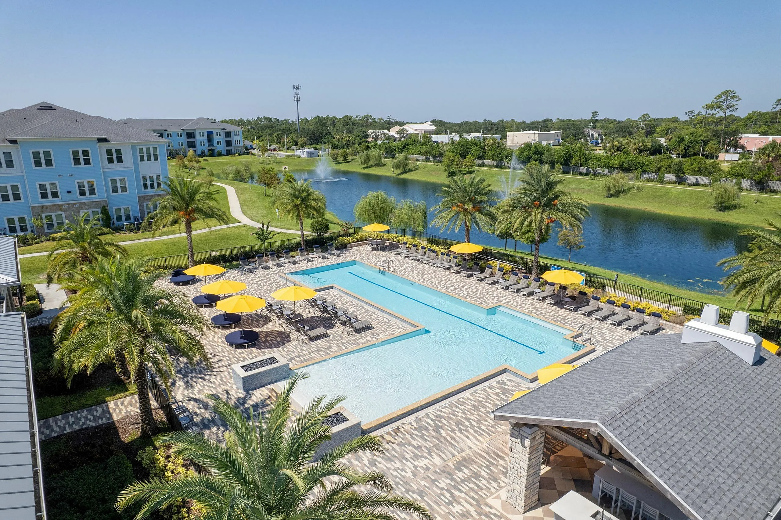 Luxury apartment complex with beautiful palm-lined pool area, vibrant yellow umbrellas, and scenic lake view, offering resort-style living.
