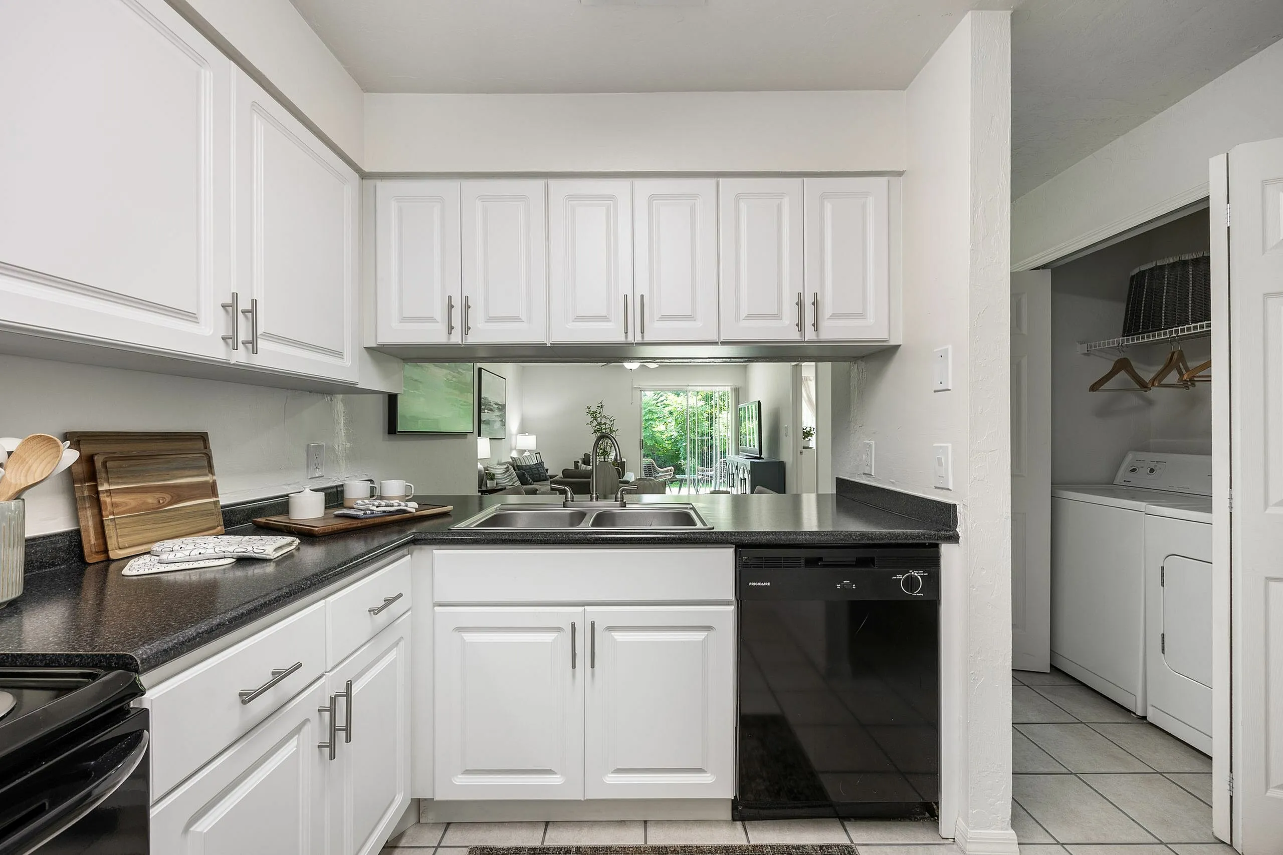 Contemporary kitchen with sleek white cabinets and black countertops, leading into a bright living area with lush garden views.