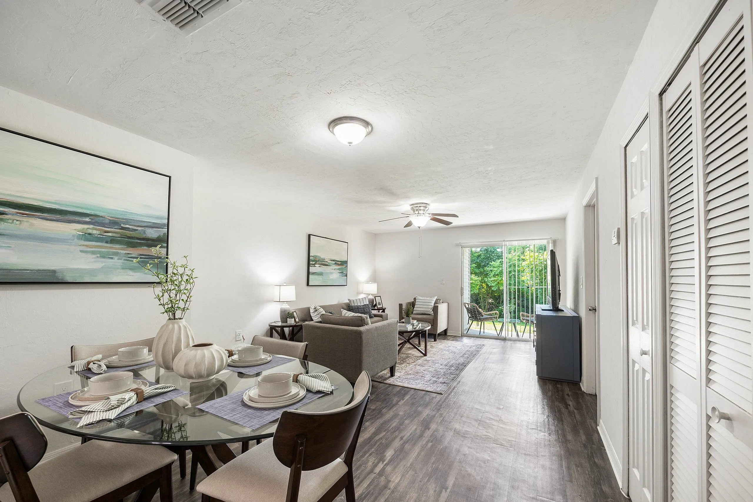 Bright and elegant apartment interior with open-plan living and dining area, featuring modern furniture, a stylish glass dining table, and a view of the lush green garden through large sliding doors.