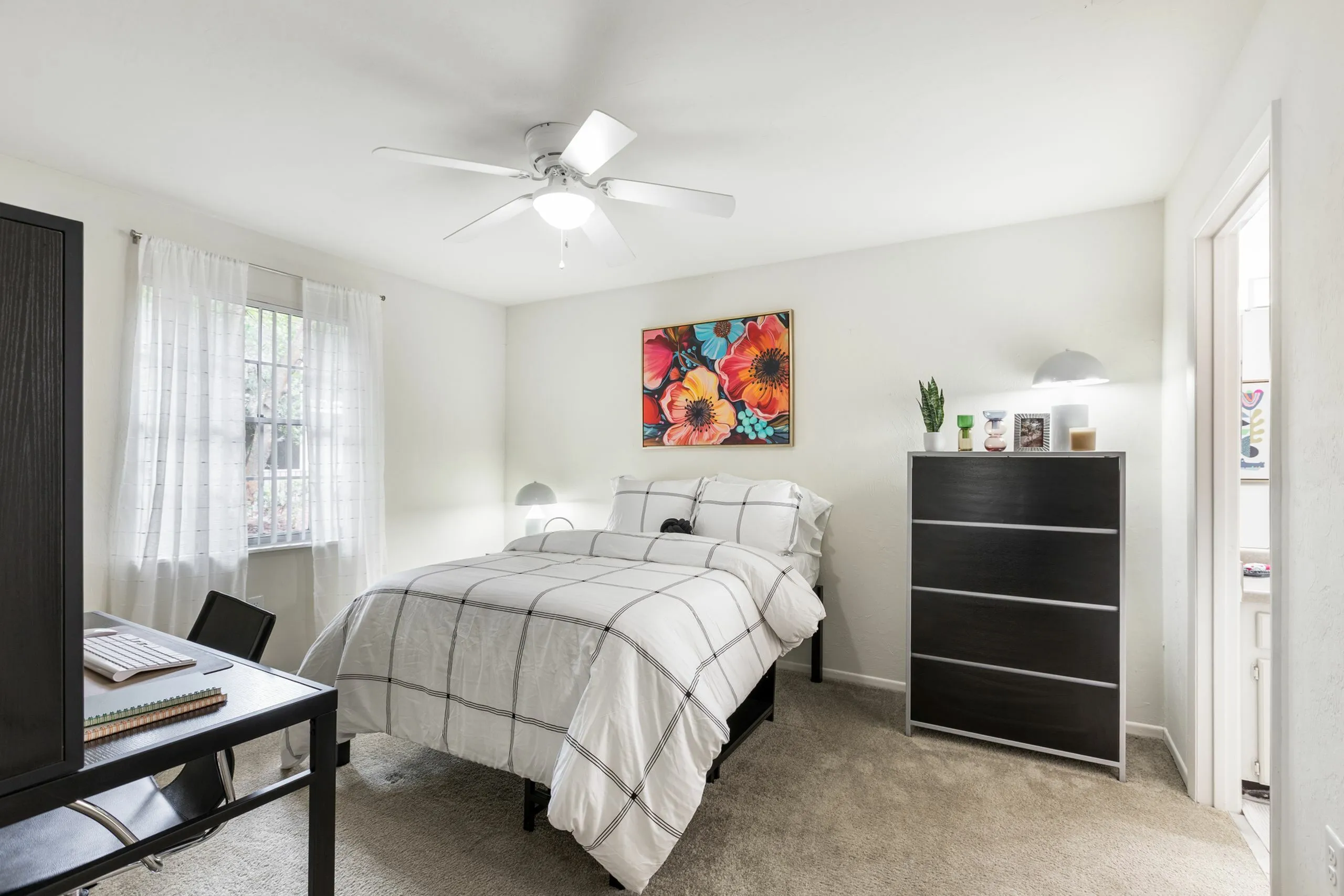 Cozy bedroom with a stylish design, featuring a comfortable bed, large window with sheer curtains, vibrant floral wall art, and sleek black furniture, creating a peaceful atmosphere.