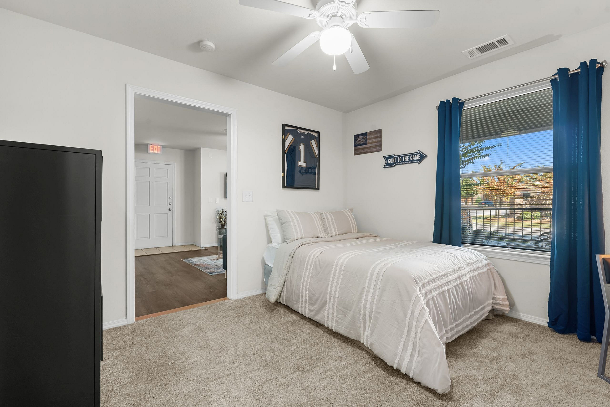 Bright and inviting bedroom with plush carpeting, elegant bed, sporty decor, and sunlit window with blue curtains.