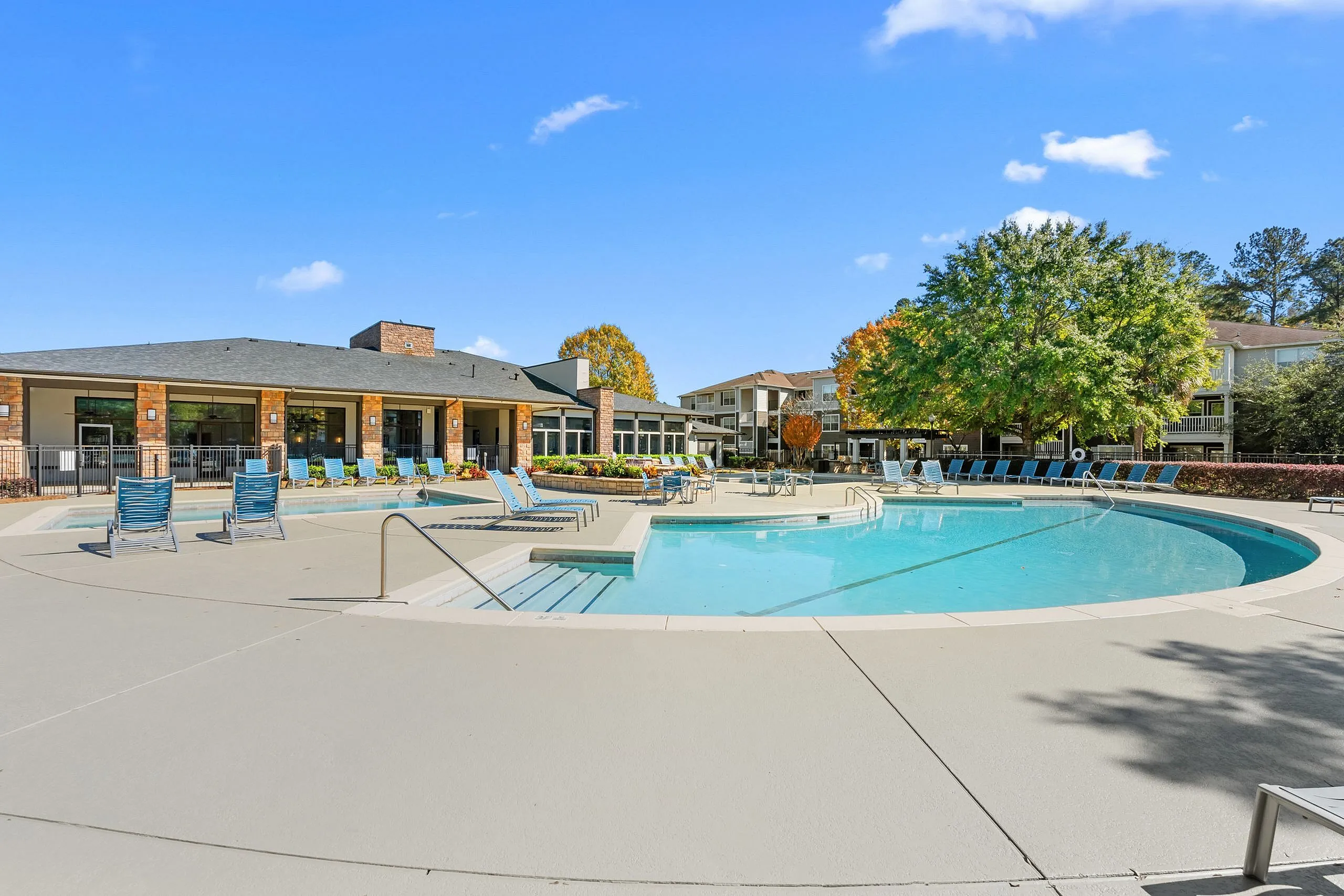 Luxurious outdoor pool area with modern lounge chairs, surrounded by vibrant landscaping and a clubhouse under a clear blue sky.