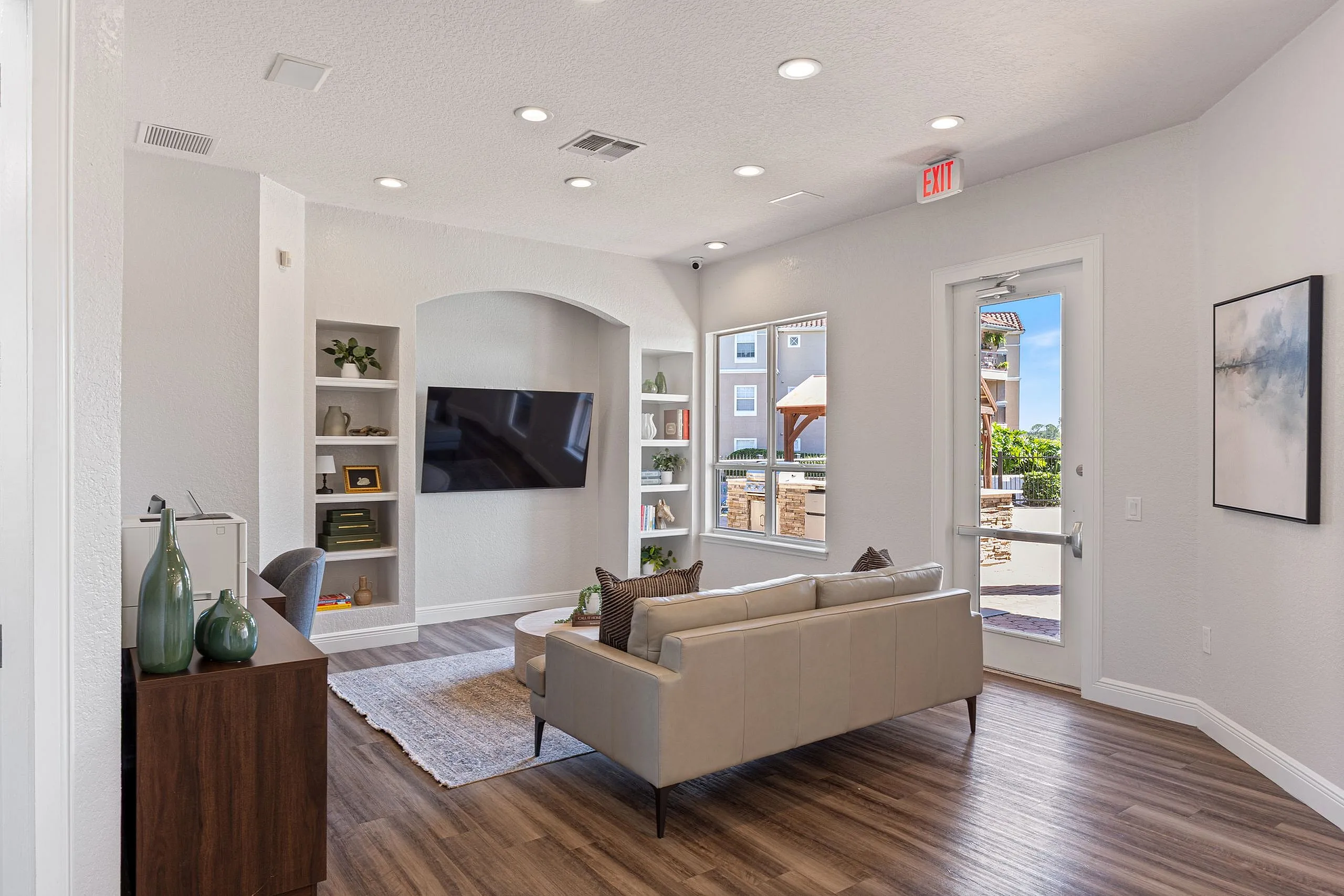 Bright and modern living room with elegant decor, featuring a stylish leather sofa, built-in shelves, and a large flat-screen TV, all set against a backdrop of sleek hardwood flooring and abundant natural light from large windows.