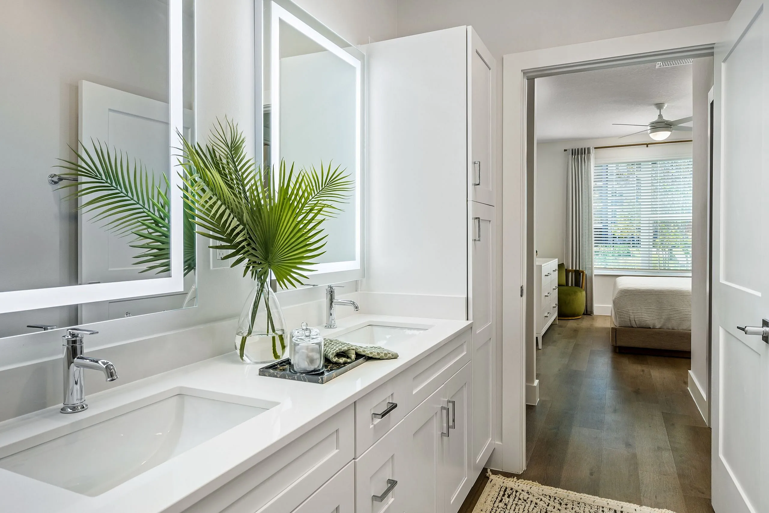 Modern bathroom with elegant double vanity, illuminated mirrors, and a view into a cozy bedroom, accentuated by natural light through large windows.