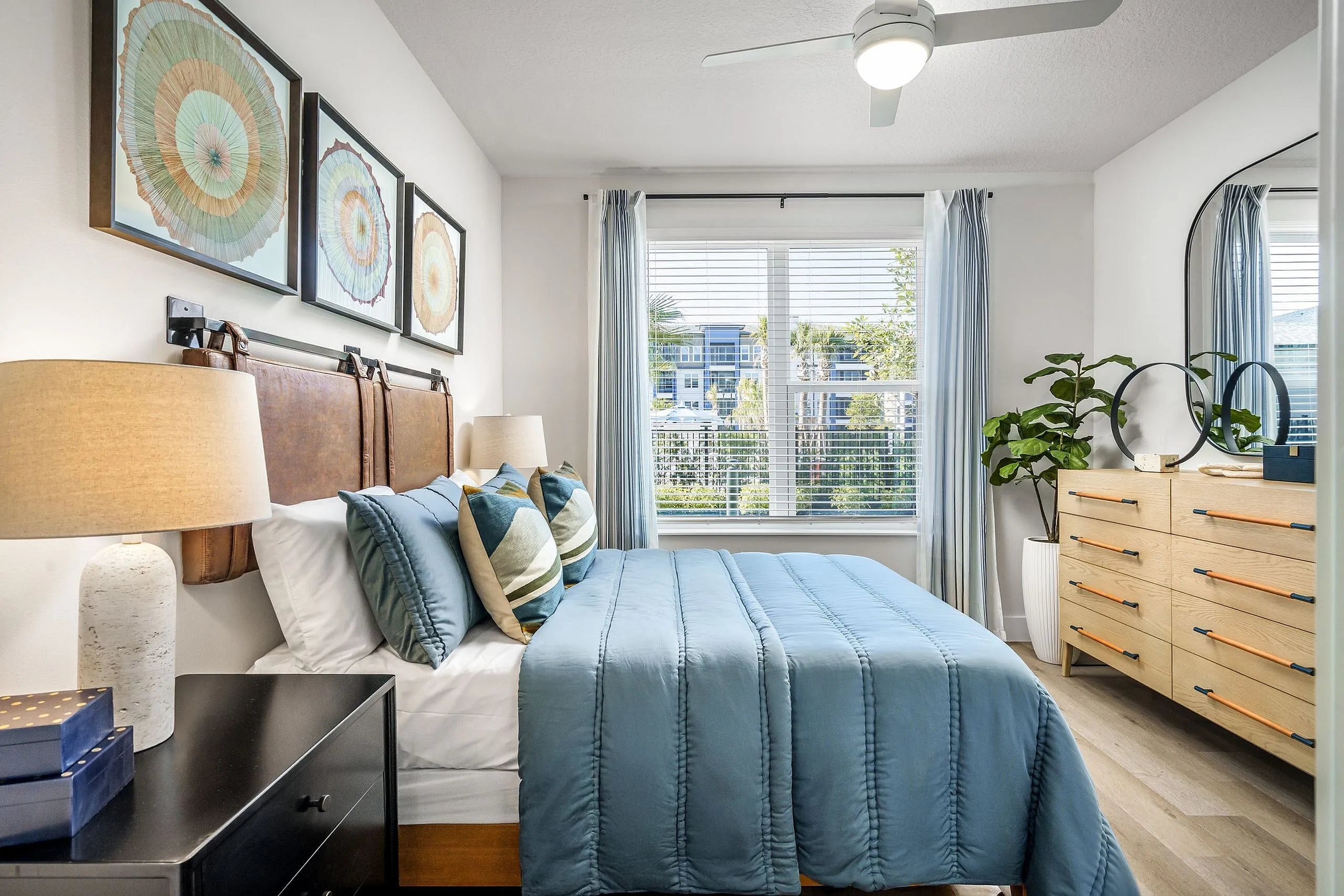 Inviting bedroom with stylish decor featuring a comfortable blue bedding set, elegant artwork, and abundant natural light from large windows overlooking lush greenery.